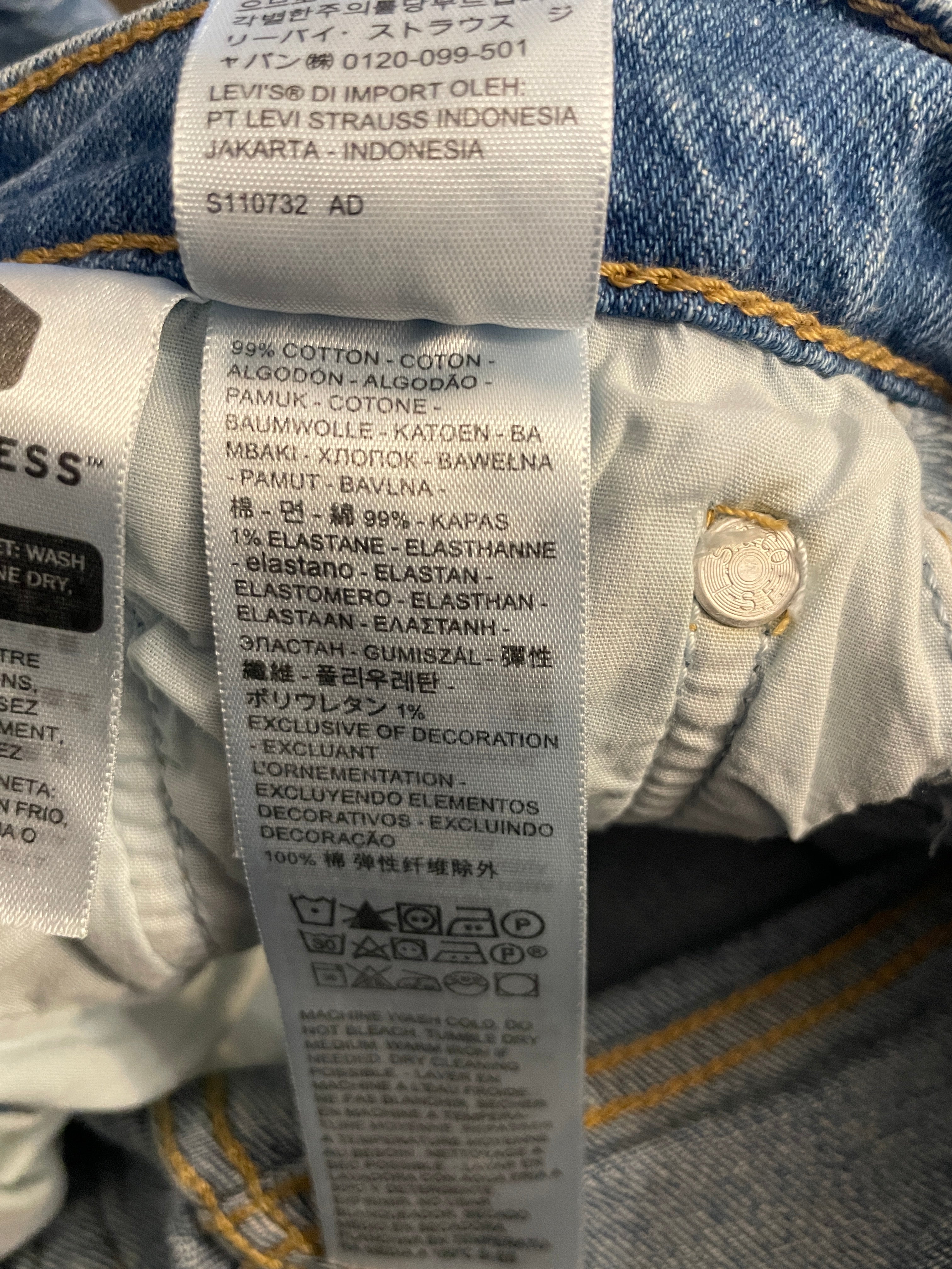 Close-up of a denim garment's inner label and hem area, showing the brand Levi's text and care instructions in Indonesian, with blue denim fabric and a gold-tone rivet visible.