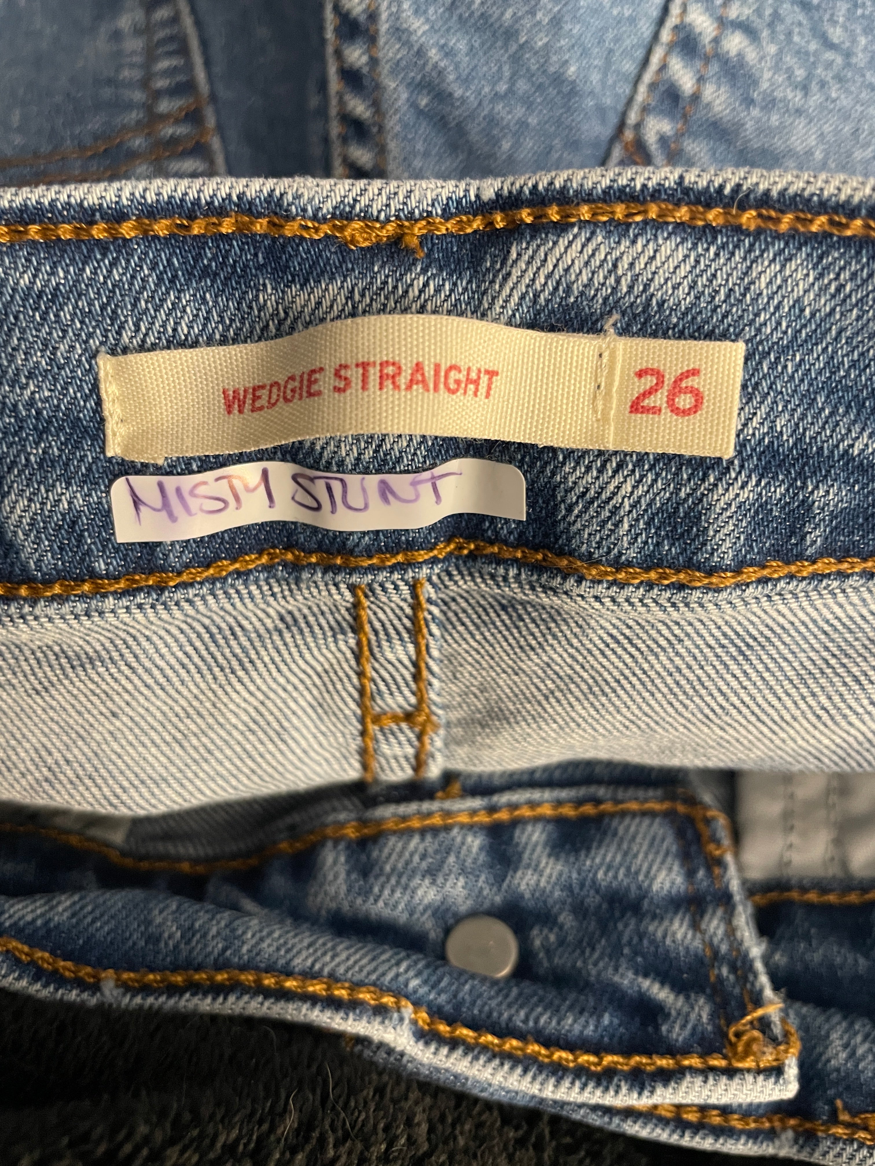 Close-up of the inside waistband of a pair of blue denim jeans, showing the size label 'WEDGIE STRAIGHT 26' and a handwritten size note that reads 'MISTY STUNT'.