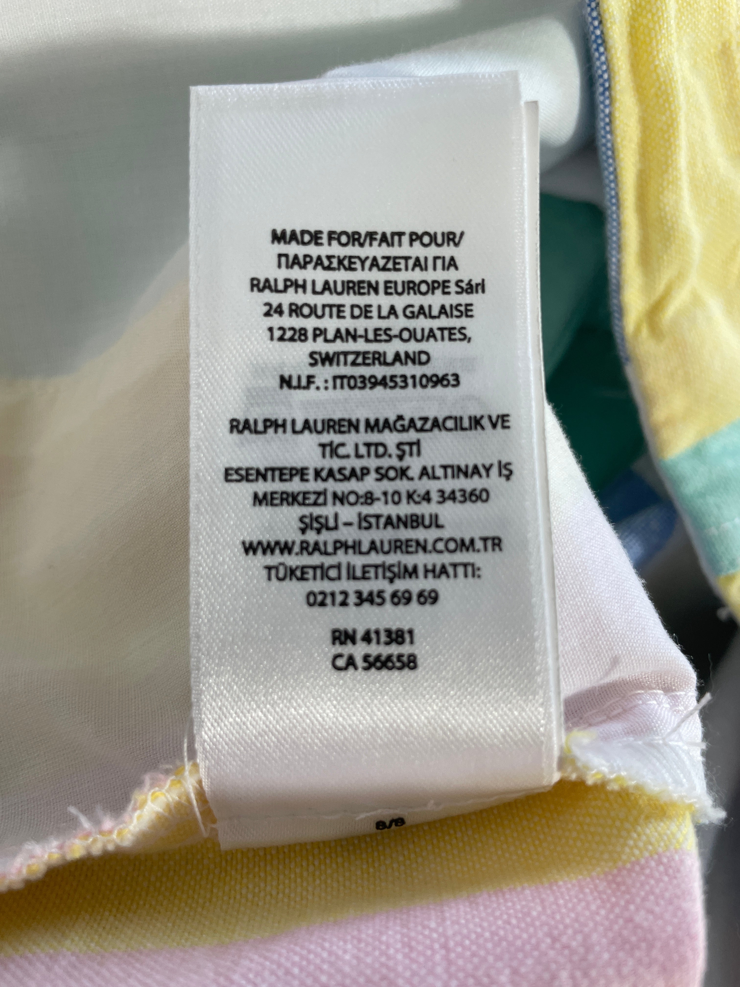 A close-up of a white fabric care label sewn onto a pastel multicolor garment, showing the text 'RALPH LAUREN EUROPE Sérl' and Turkish address details.