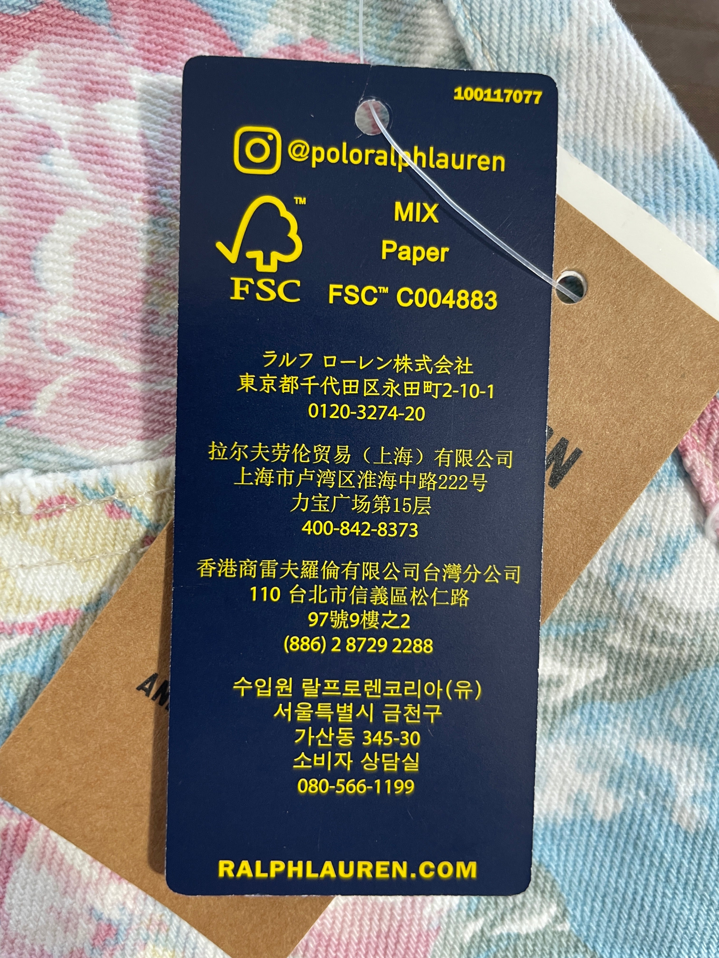 A dark navy blue Ralph Lauren hangtag with yellow text and icons, photographed over a kraft paper tag on a pink and blue patterned fabric background.