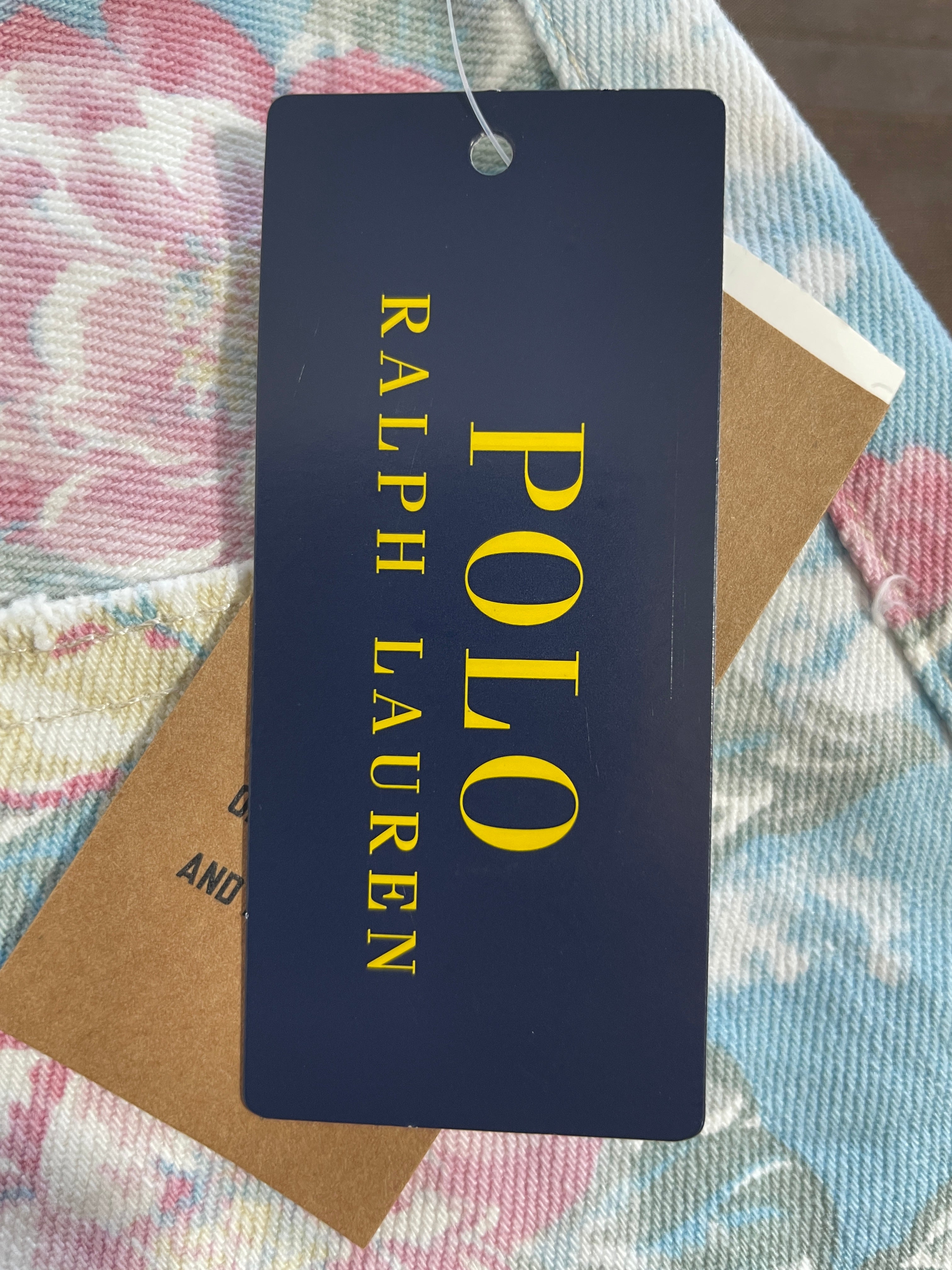 A navy blue Ralph Lauren Polo tag with yellow lettering, attached to a light blue and pink floral-patterned garment, with a brown kraft paper tag underneath.