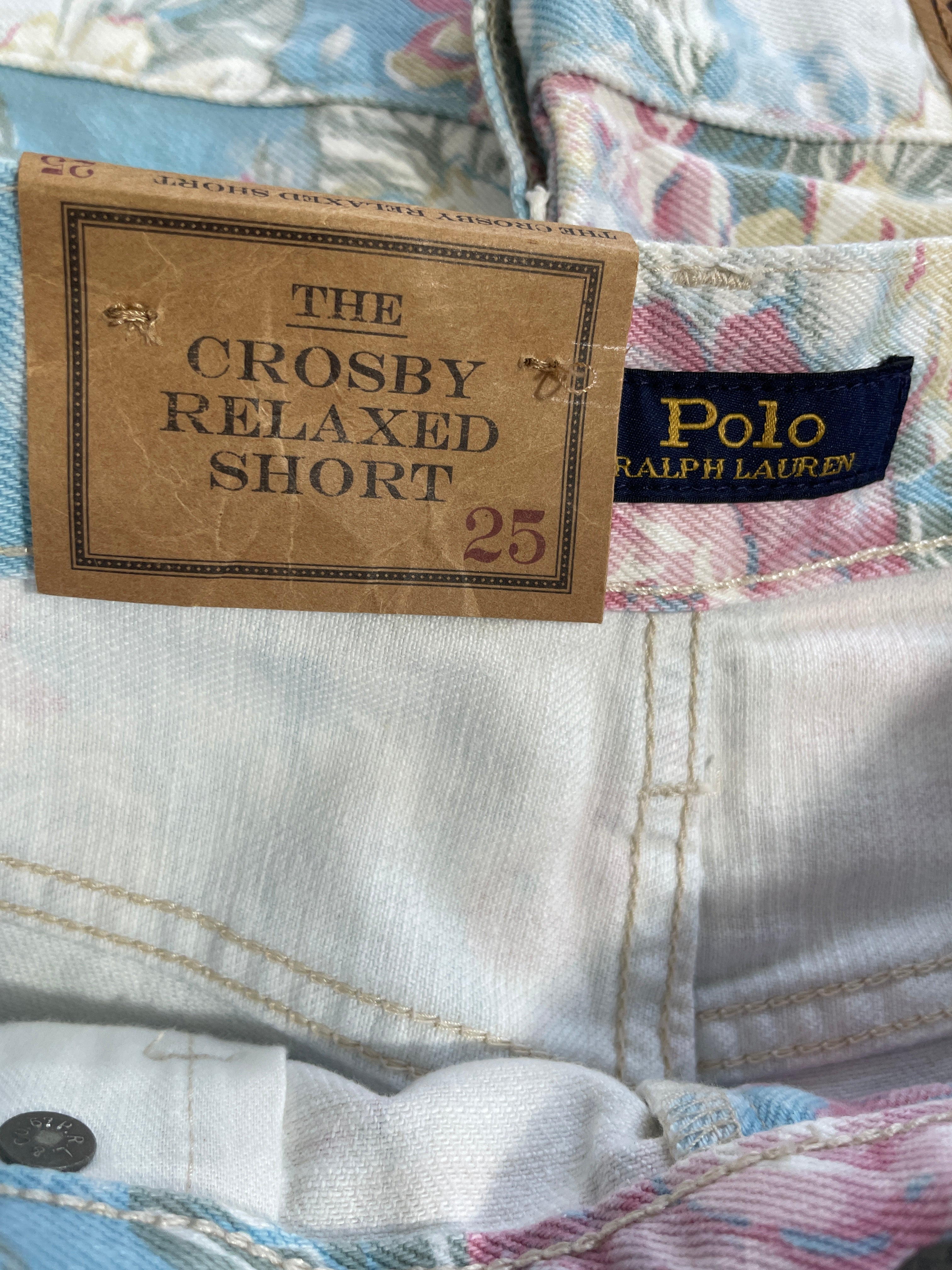 Close-up of a Polo Ralph Lauren denim short with a brown paper hangtag reading 'THE CROSBY RELAXED SHORT 25' and a navy Polo Ralph Lauren neck label.