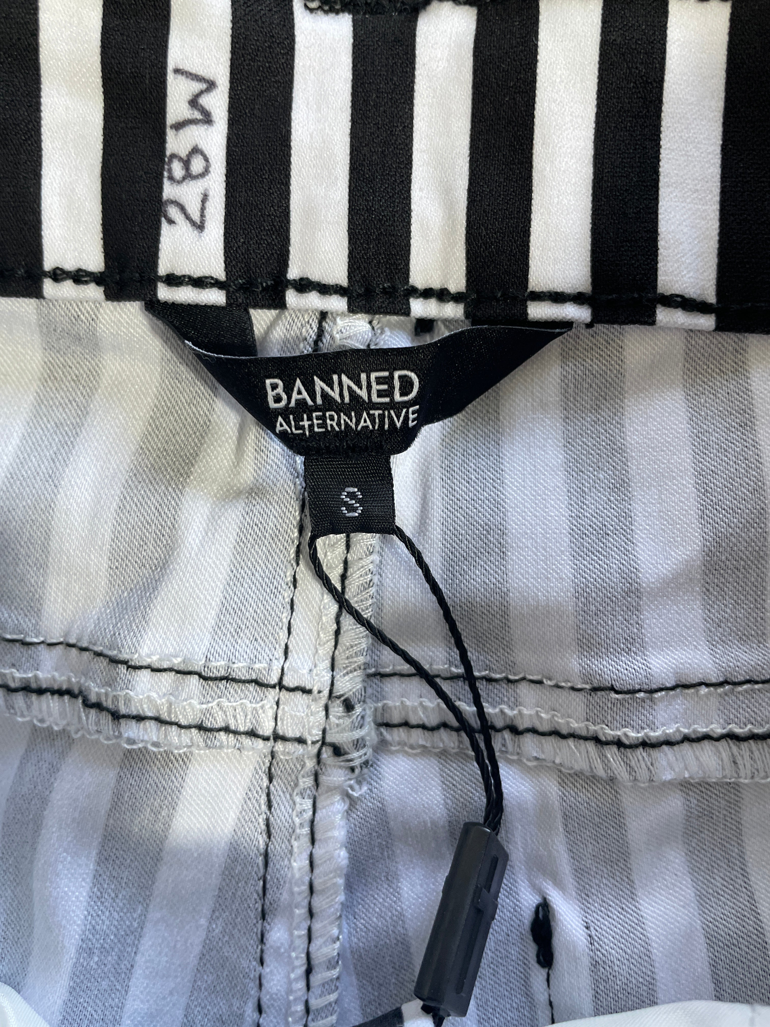 Close-up of striped pants fabric with a black BANNED ALTERNATIVE brand label and a black hangtag attached, showing a white, gray, and black stripe pattern.