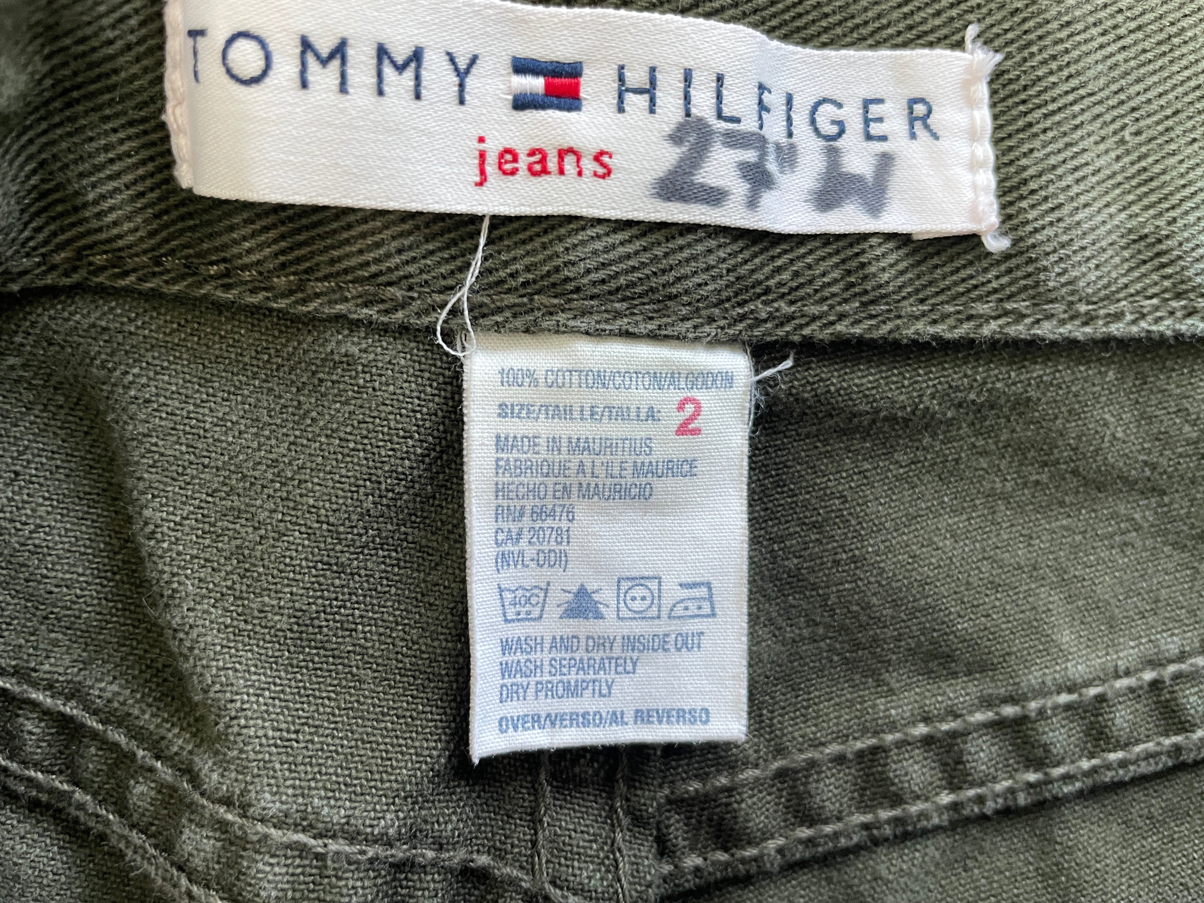 Close-up of a Tommy Hilfiger jeans label showing the brand name, 'jeans' text, and a size '2' care tag stitched onto the inside waistband of an olive green garment.