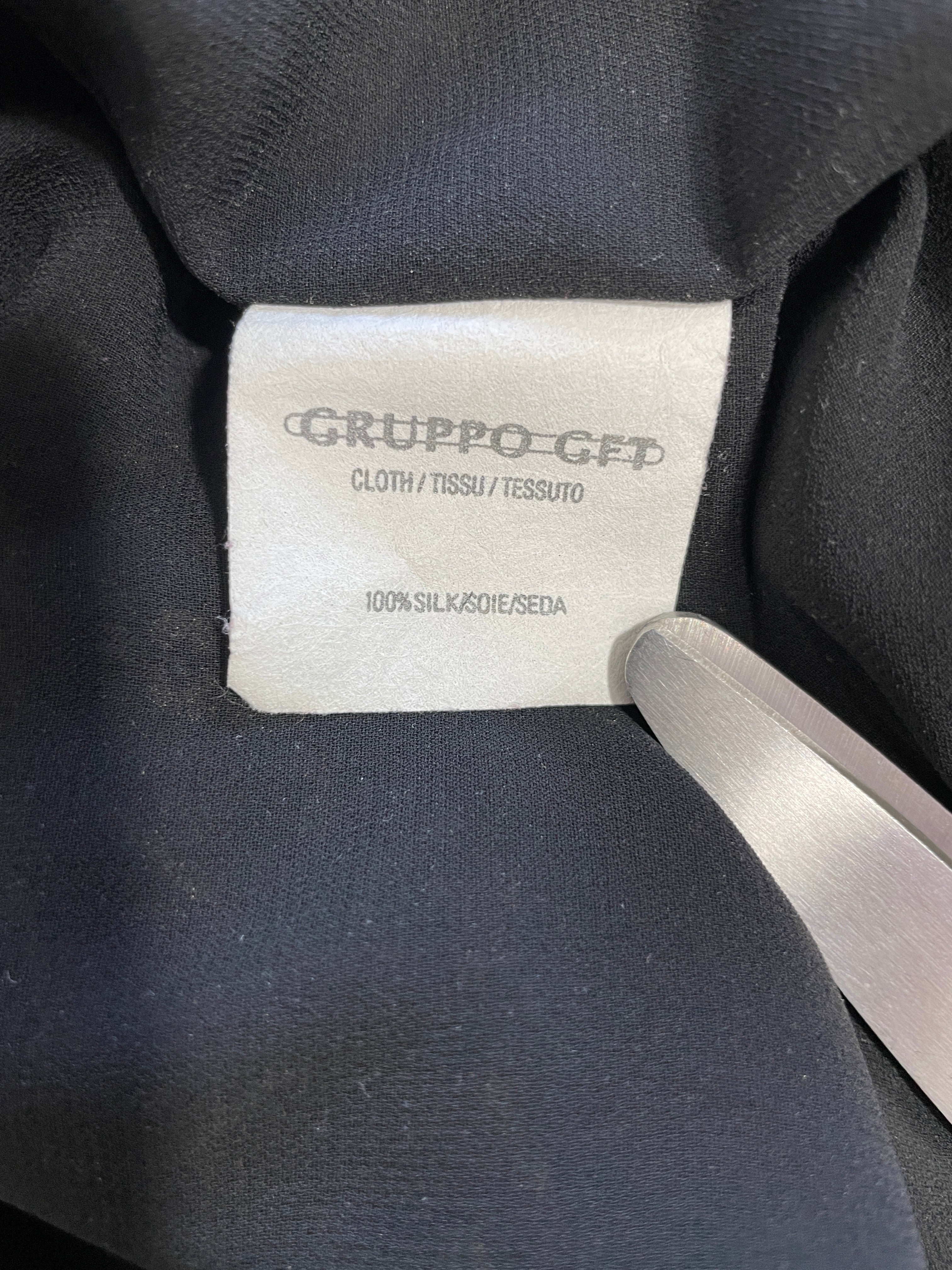 Close-up of a clothing label reading GRUPPO-GET with 'CLOTH / TISSUTO / TESSUTO' and '100% SILK/SEIDE/SEIDA' on dark navy fabric, with a metal tool touching the label.