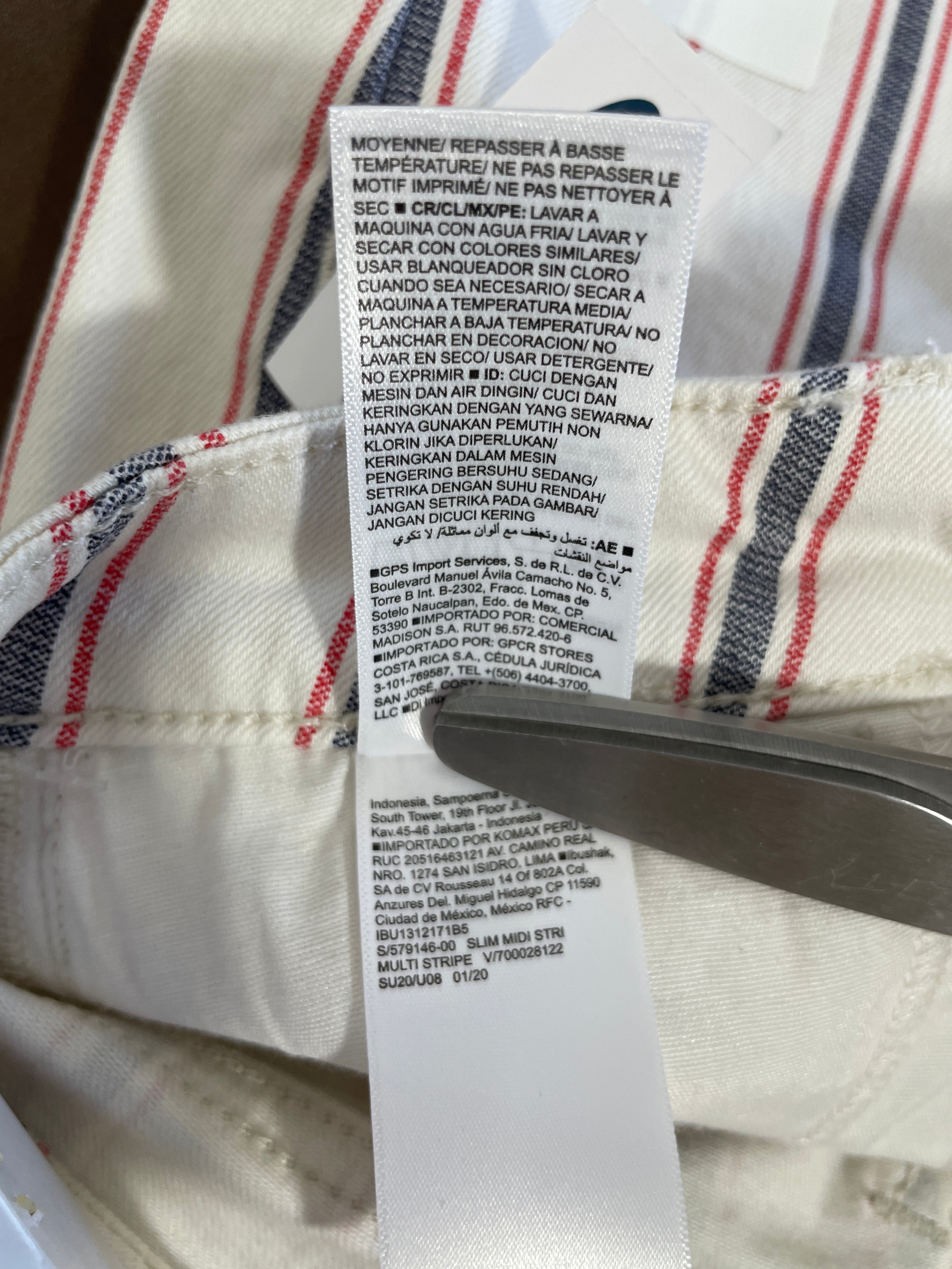 Close-up of a garment's care label sewn onto an off-white fabric with red and navy stripes, with a metal tool holding the label open.