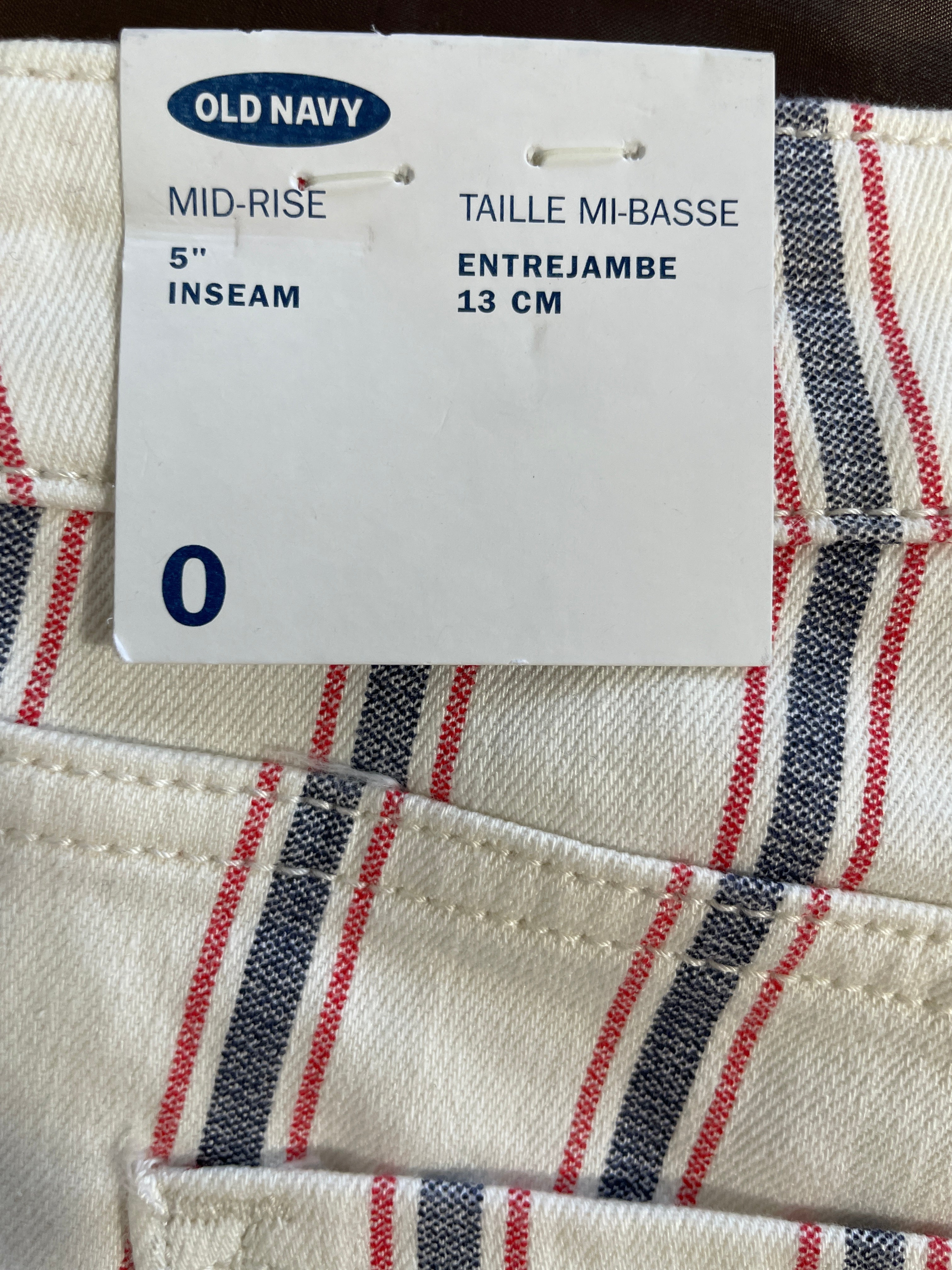 Close-up of striped fabric with a white Old Navy size tag attached. The textile is off-white with navy and red diagonal stripes, and the tag reads 'TAILLE MI-BASSE ENTREJAMBE 13 CM', '5" INSEAM', and 'OLD NAVY'.