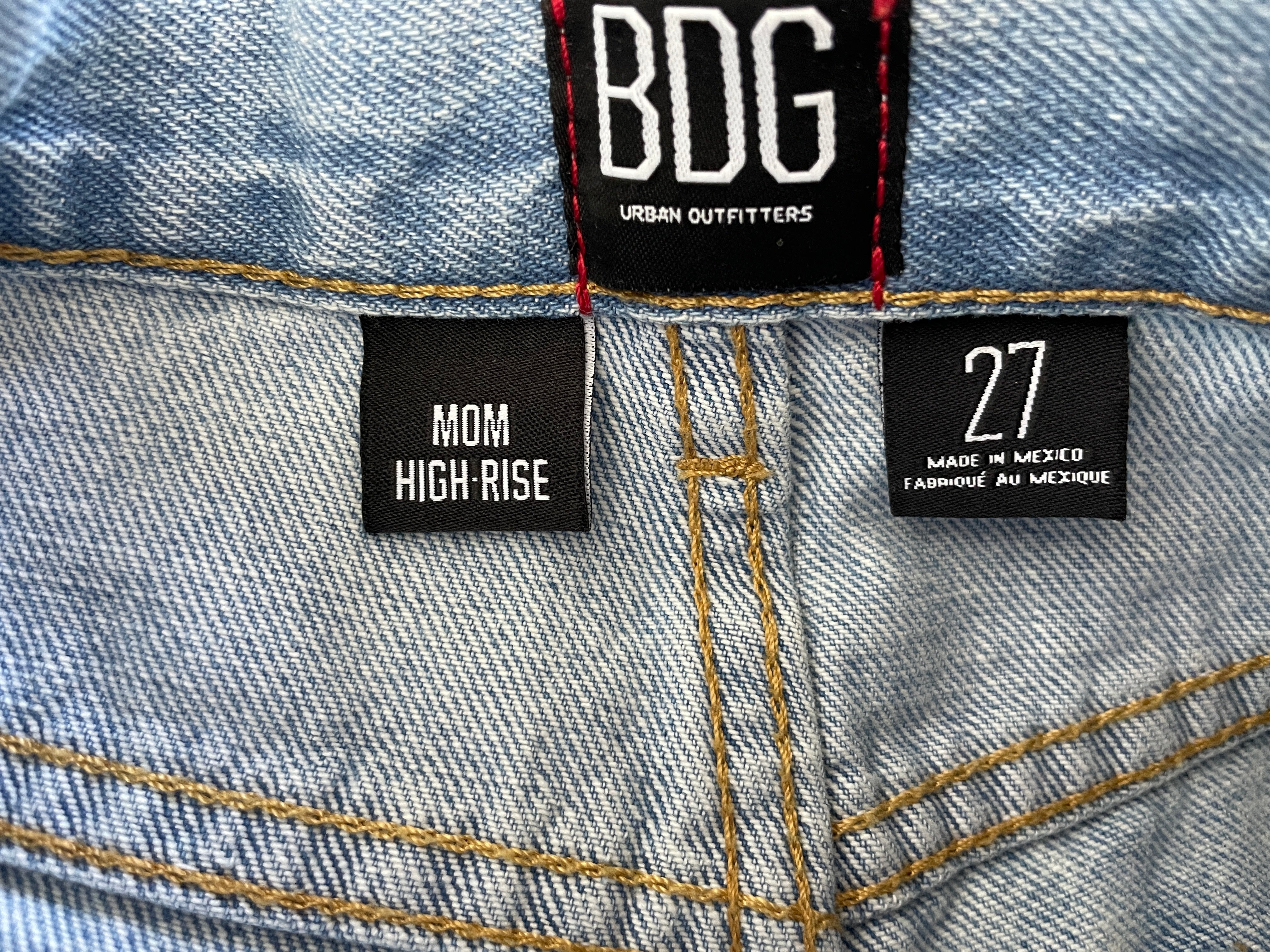 Close-up of the back waistband of light-wash blue denim jeans showing the brand label 'BDG URBAN OUTFITTERS', a 'MOM HIGH RISE' size label, and a '27 MADE IN MEXICO FASHION IN MEXICO' label.