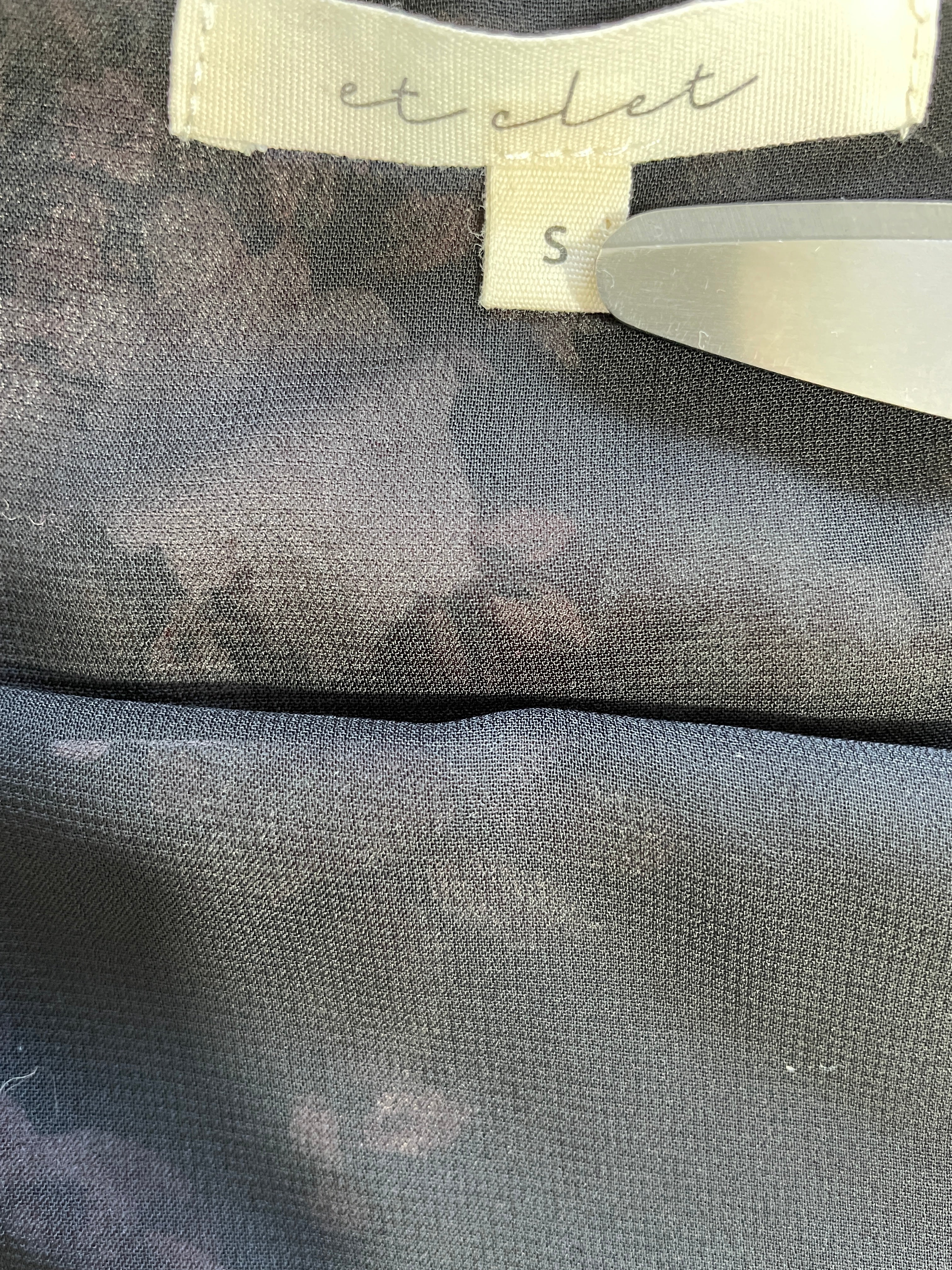 Close-up of a garment label reading 'et olet' with size 'S' on a dark gray patterned fabric, with a plastic tag applicator visible on the right.