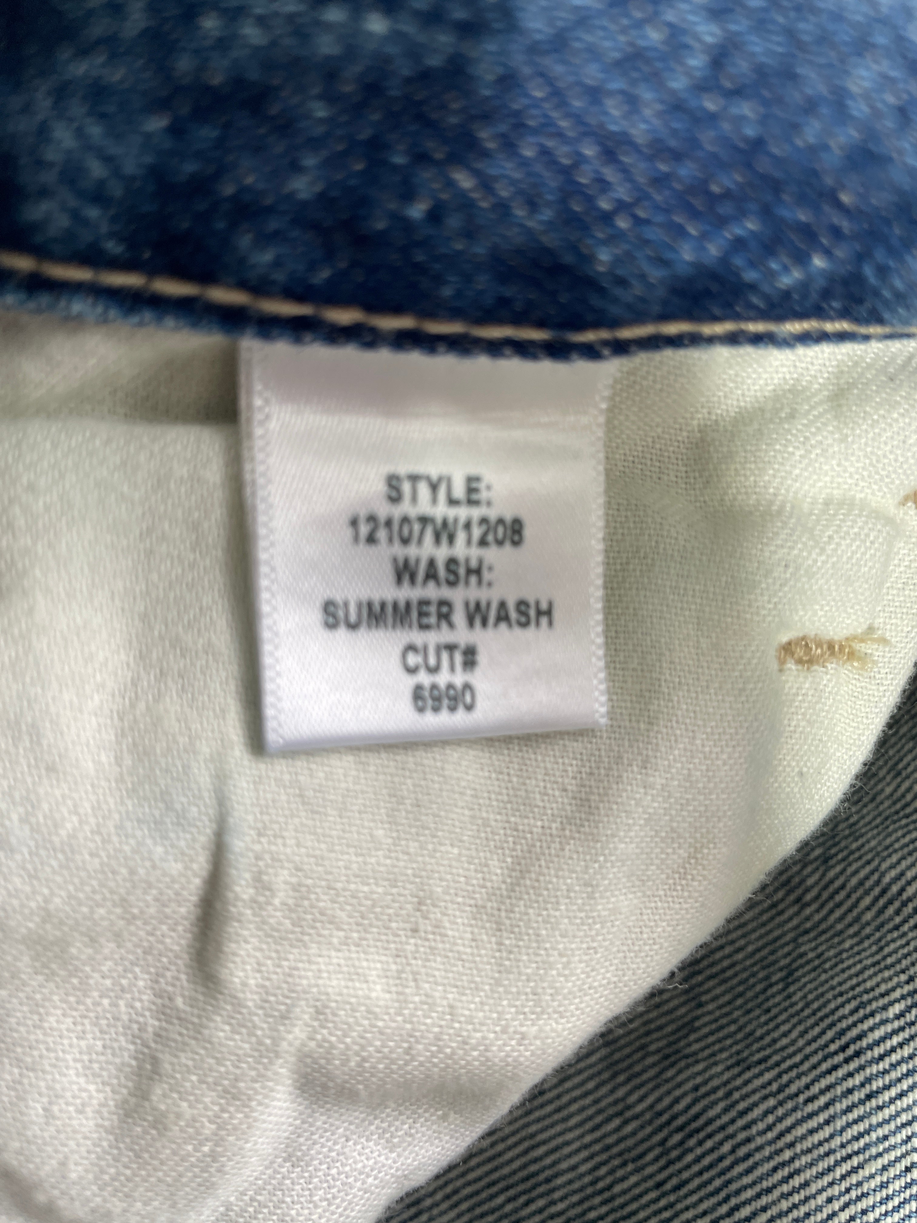 Close-up of a garment's interior neck label reading 'STYLE: 12107W1208 WASH: SUMMER WASH CUT#: 6990' attached to a light gray fabric, with blue denim-like trim visible at the top and bottom edges.