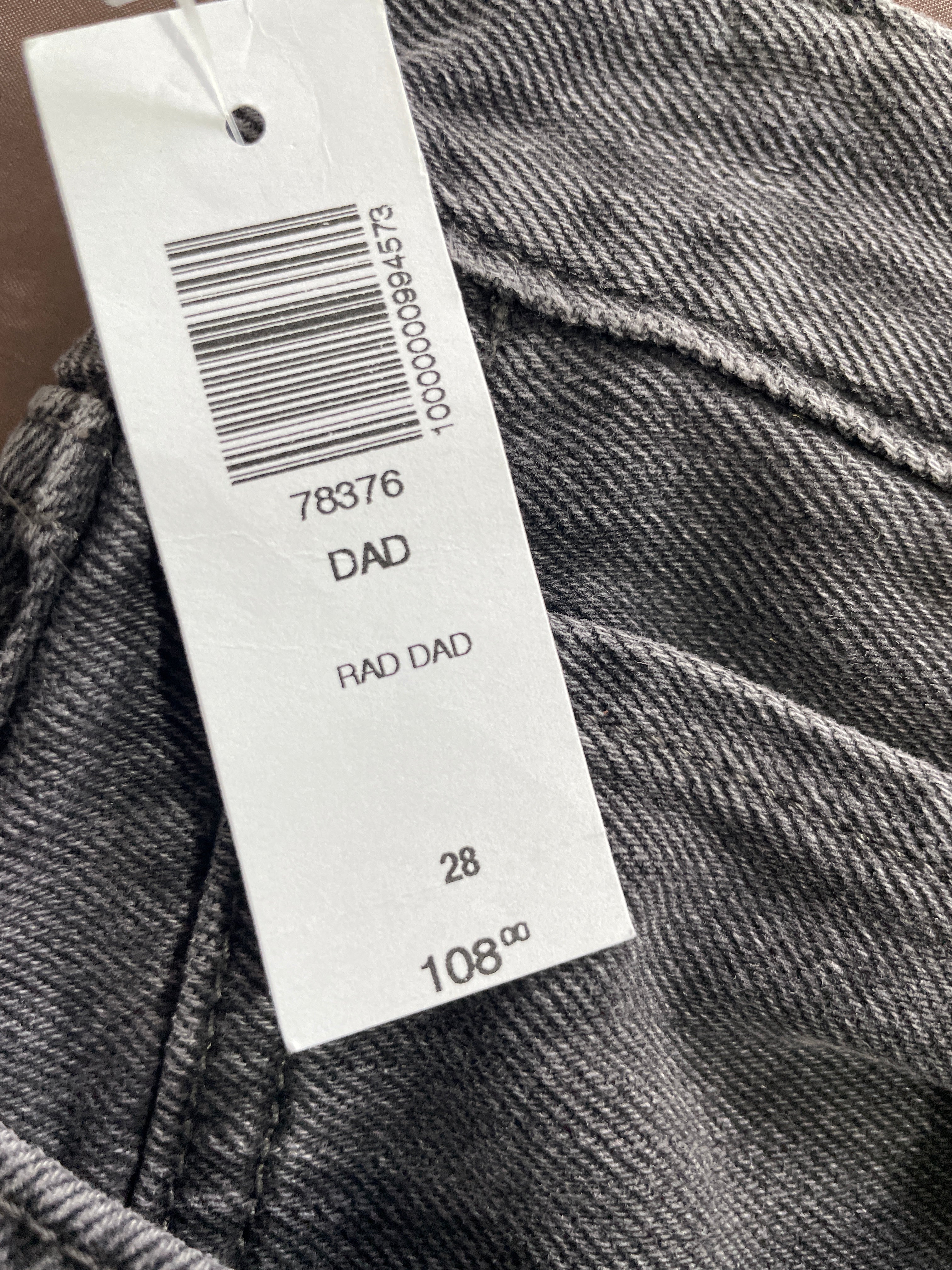 Close-up of dark gray denim fabric with a white paper size tag attached, showing printed text including 'DAD', 'RAD DAD', size '28', and price '108.99'.