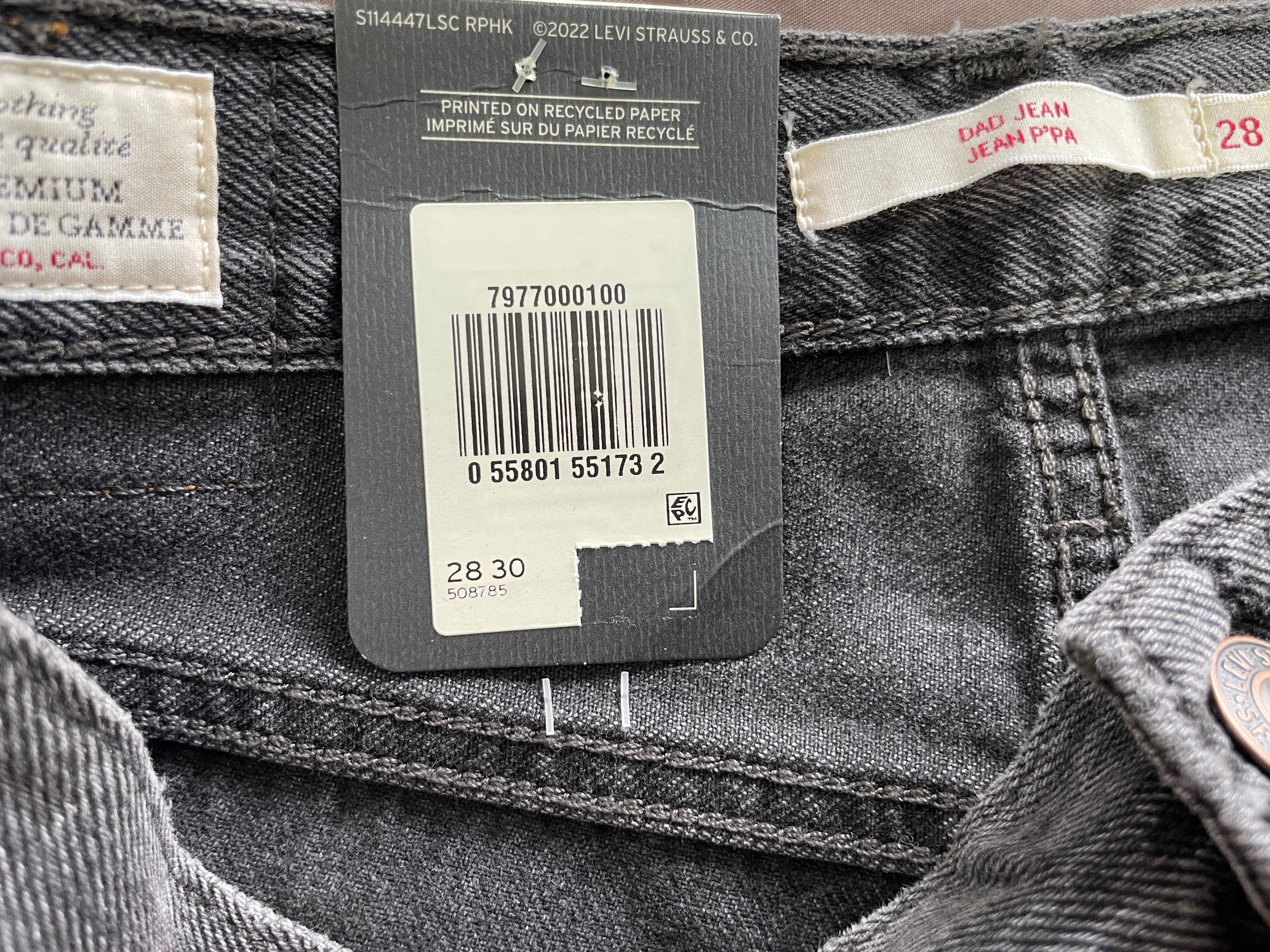 Close-up of dark gray denim jeans showing the waistband, stitching, and attached retail tags including a barcode label and a cream-colored brand tag.