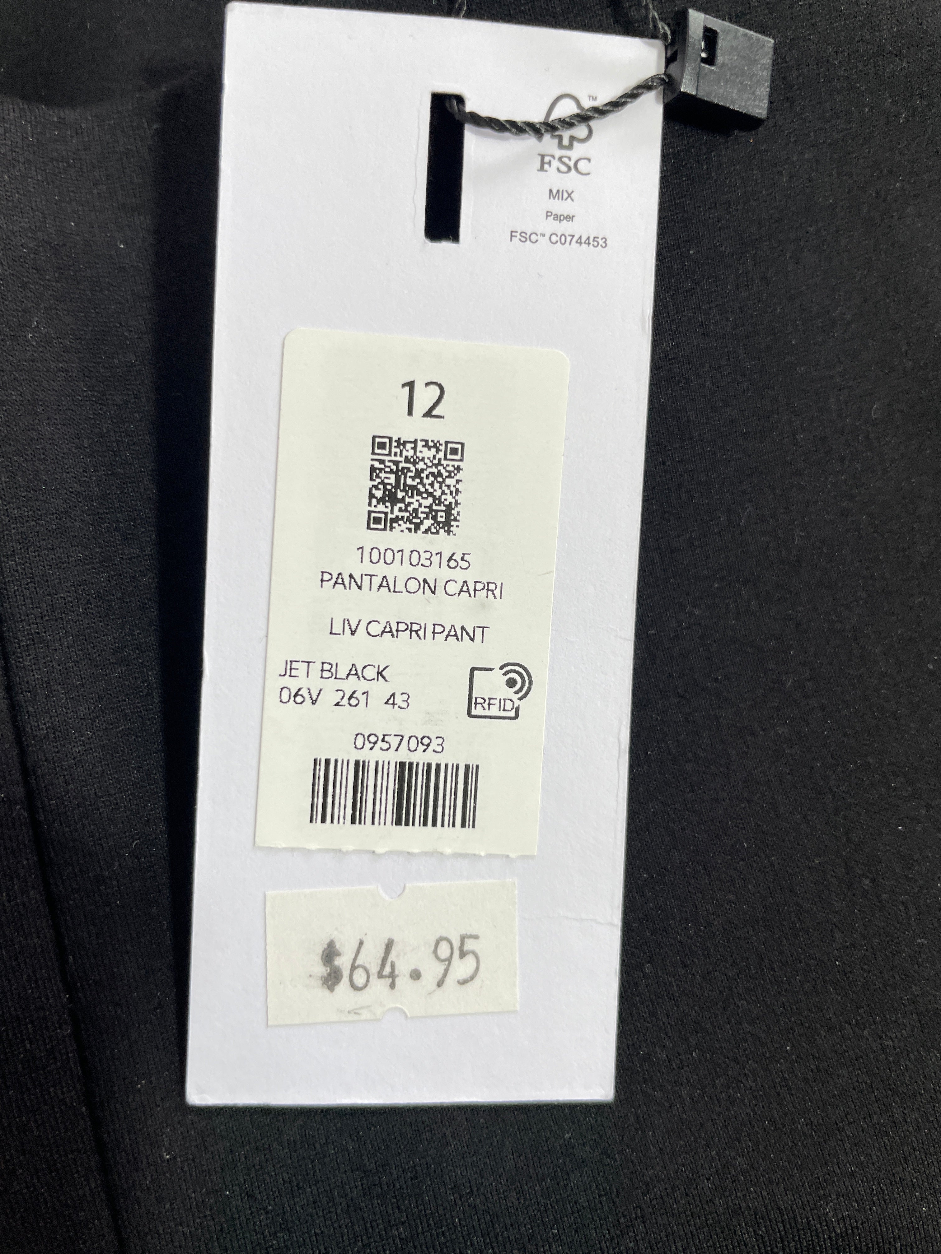 A close-up of a garment's hangtag attached to a black fabric surface. The tag shows FSC branding, size 12, product text 'PANTALON CAFRI PANT', color 'JET BLACK', and a handwritten price sticker reading $64.