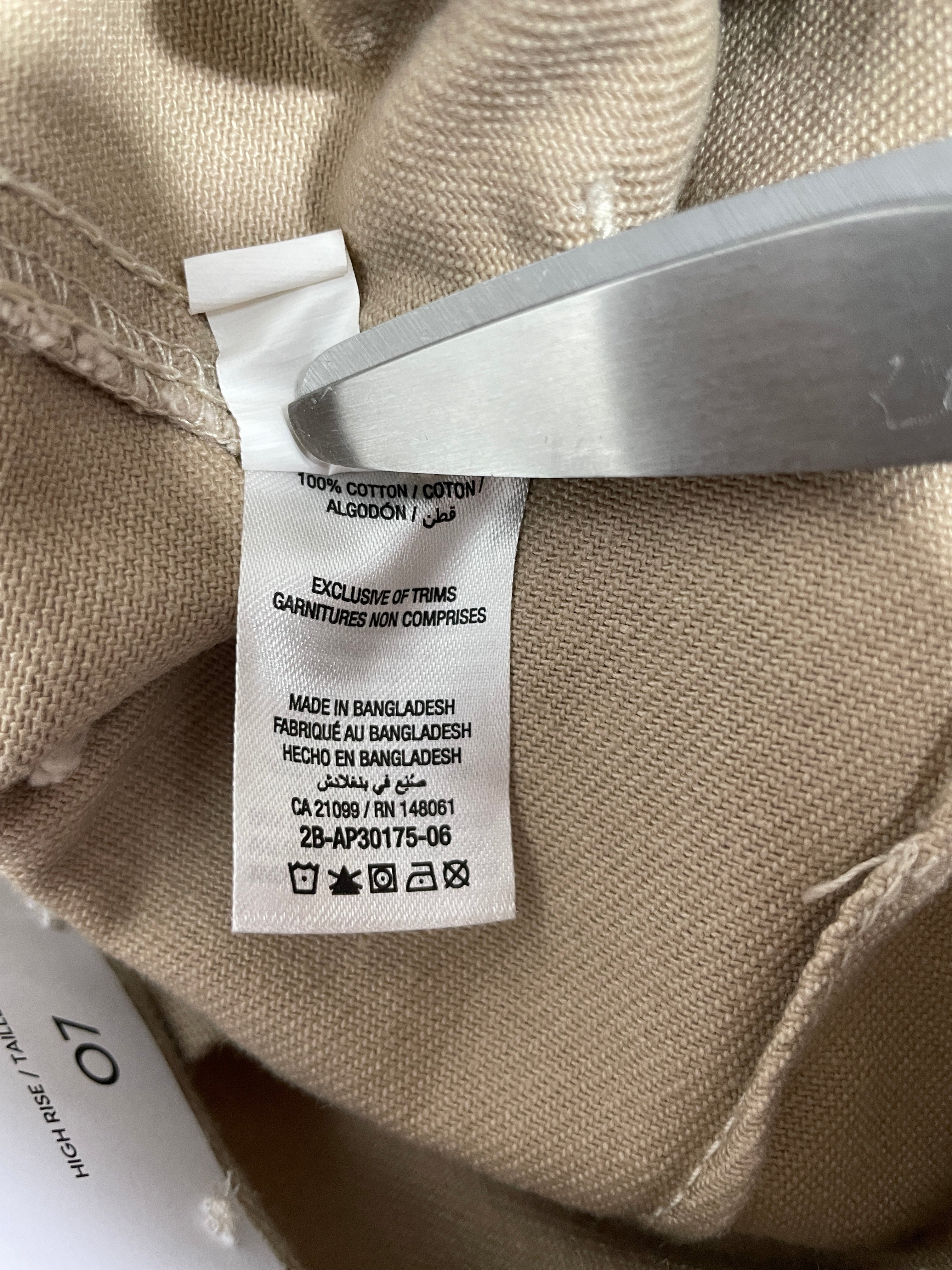 Close-up of a beige garment's interior label sewn onto the inside neck, with a metal tool holding the label in place. The fabric is a woven beige material, and a white paper tag is partially visible at the bottom right.