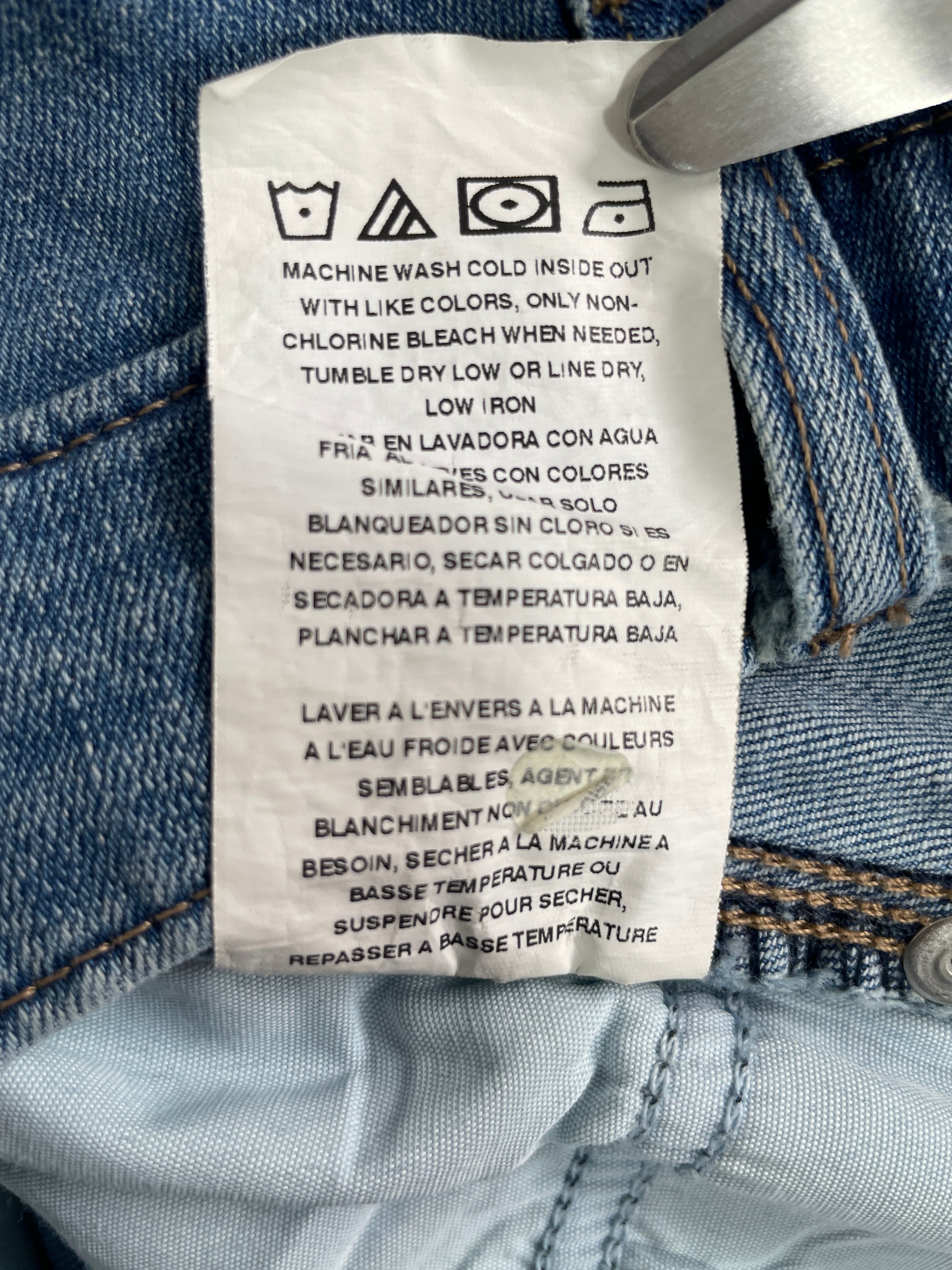 A close-up of a white garment care label sewn onto light blue denim jeans, showing washing instructions in English, Spanish, and French.
