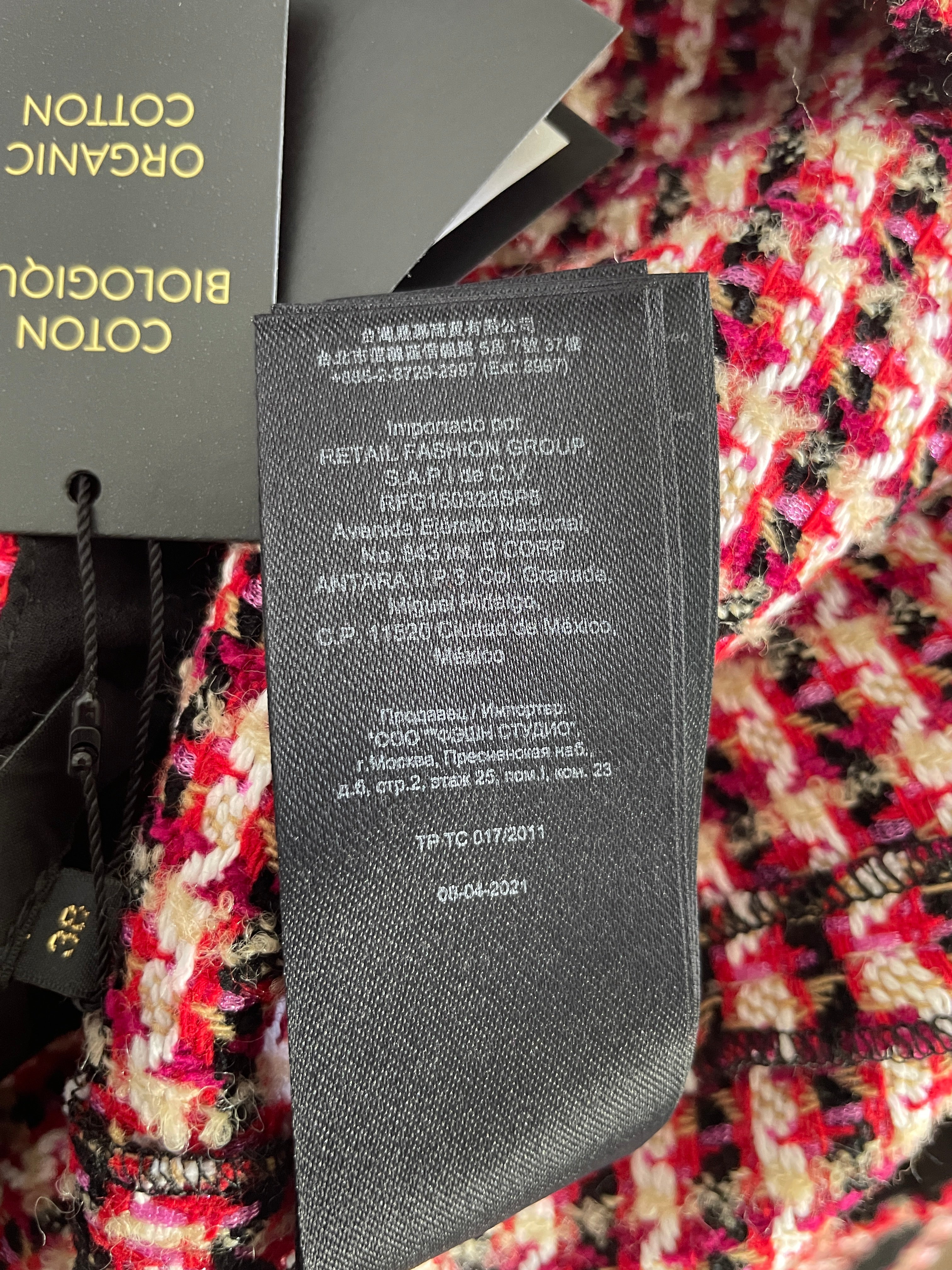 Close-up of a black fabric care tag attached to a garment, showing text including 'COTON BIOLOGICO' and 'Importado por RETAIL FASHION GROUP', with part of a black hangtag and a red, pink, and black patterned textile visible in the background.