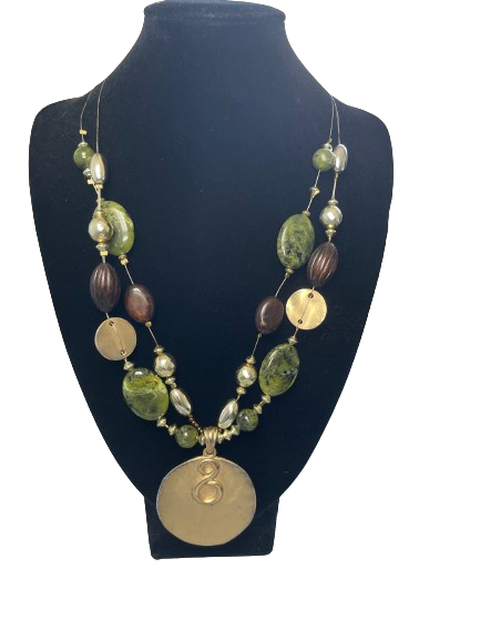 Statement necklace with green marbled beads, wood-look accents, and gold-tone discs, featuring a large round gold pendant with an infinity swirl, displayed on a black velvet stand with a wire-like double strand chain and lobster clasp.