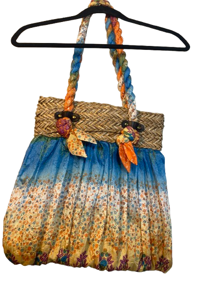 A bohemian-style shoulder bag with a braided blue, orange, and green handle, a woven straw waist strap, and a pleated skirt section featuring a blue-to-yellow floral gradient. The bag is shown hanging from a black hanger against a white background.