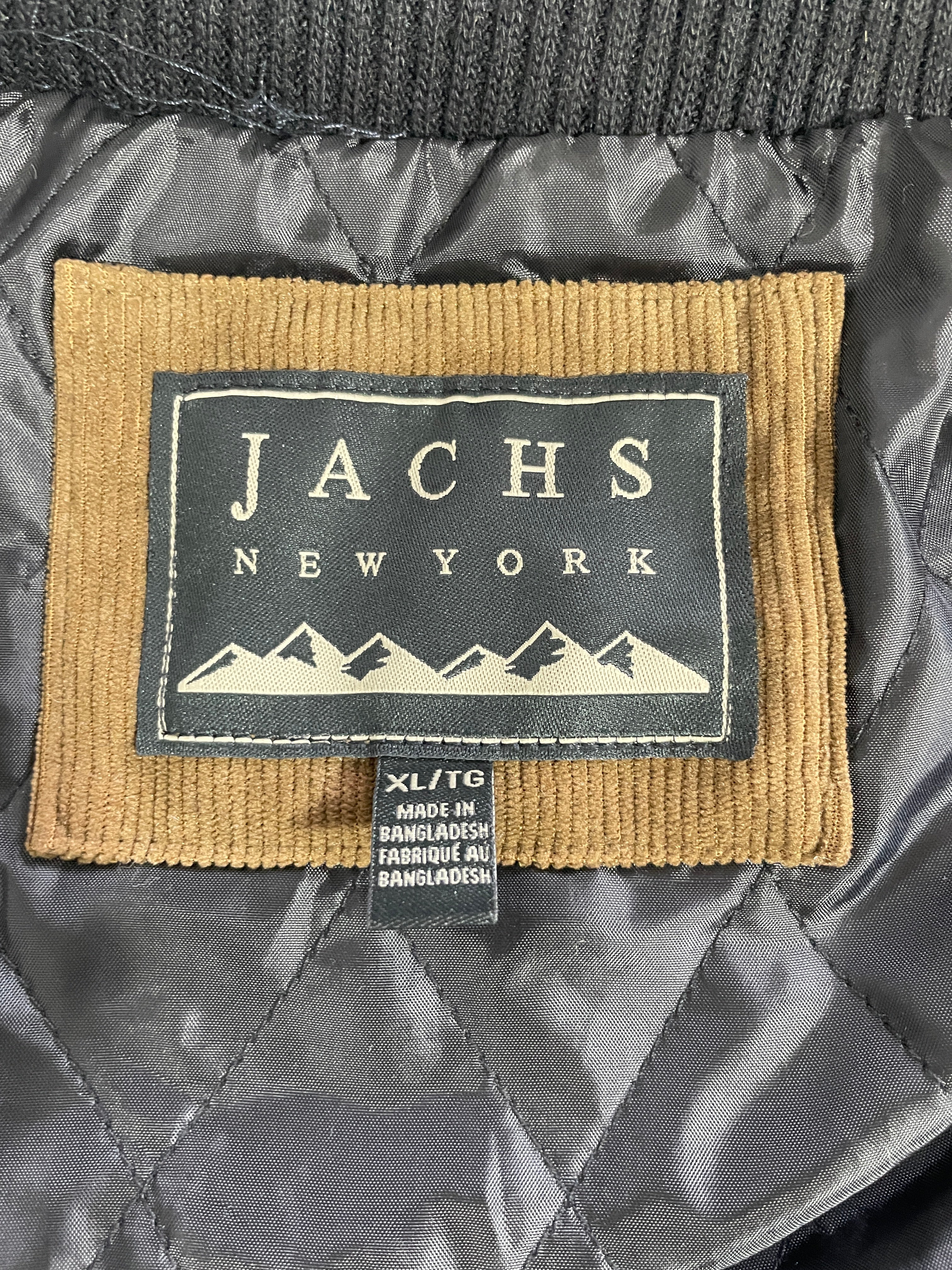 Close-up of a quilted jacket's interior label patch reading 'JACHS NEWYORK' with a mountain graphic, attached to a size tag 'XL/TG' on a dark gray fabric background.