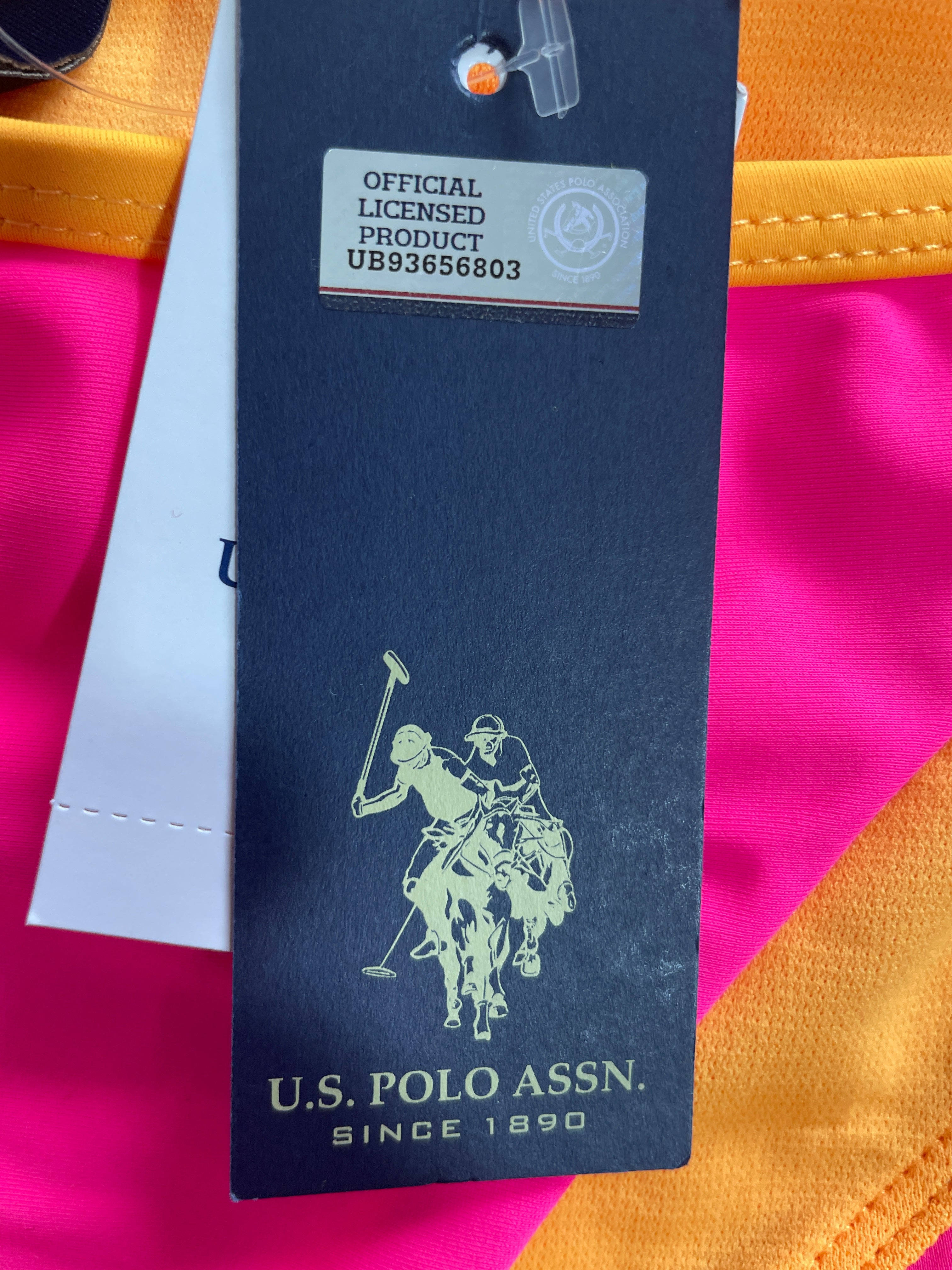 A close-up of a dark navy U.S. Polo Assn. hangtag with a gold polo player logo, placed over bright pink and orange fabric.