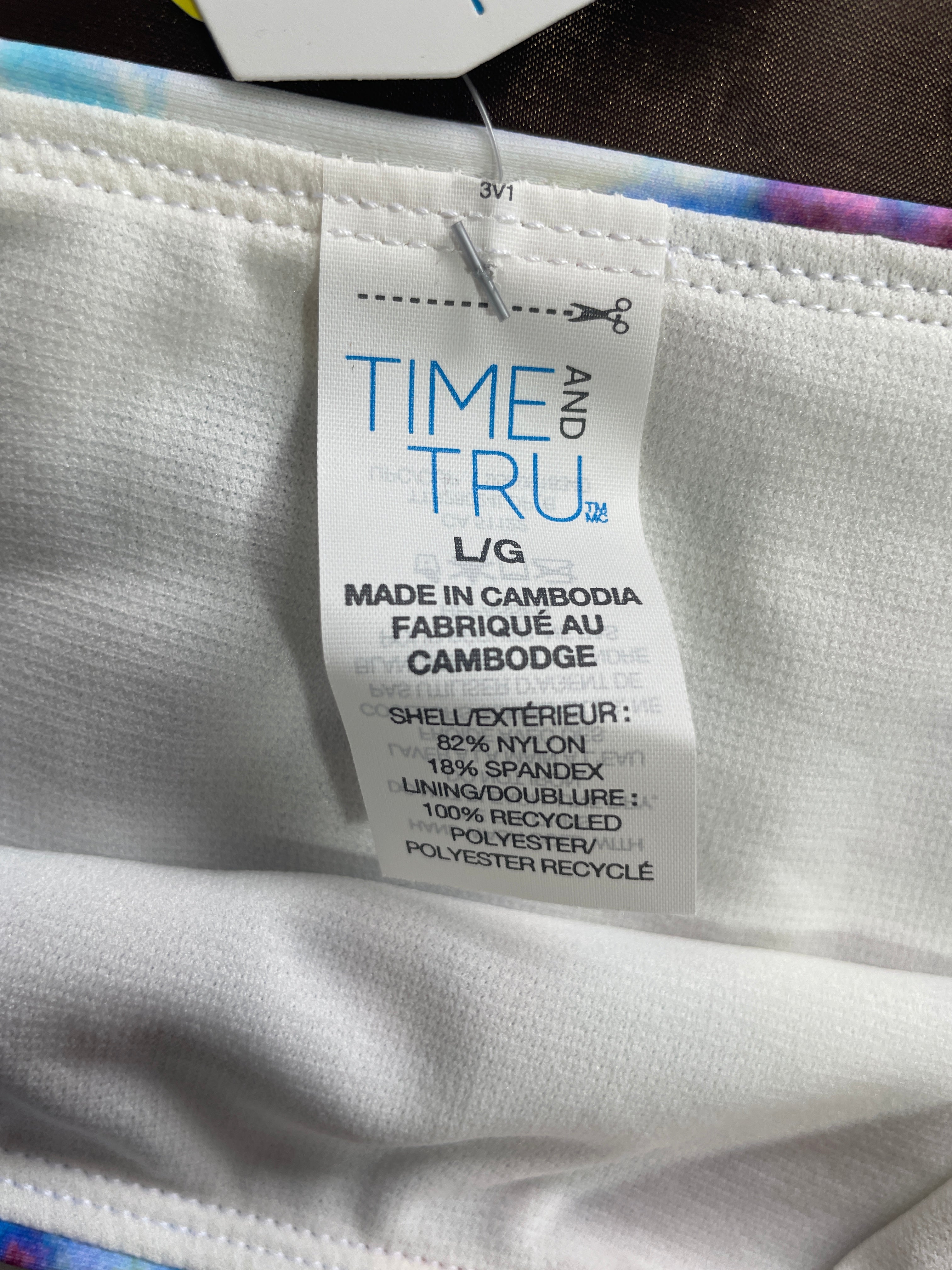 Close-up of a white garment tag reading 'TIME AND TRU' with product details including 'L/G' and 'MADE IN CAMBODIA', attached to a white ribbed fabric item.