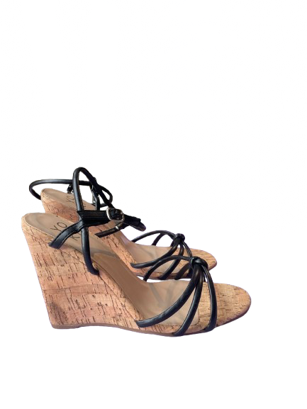 A black wedge sandal with straps and a cork wedge heel, displayed against a blurred background with Christmas lights.