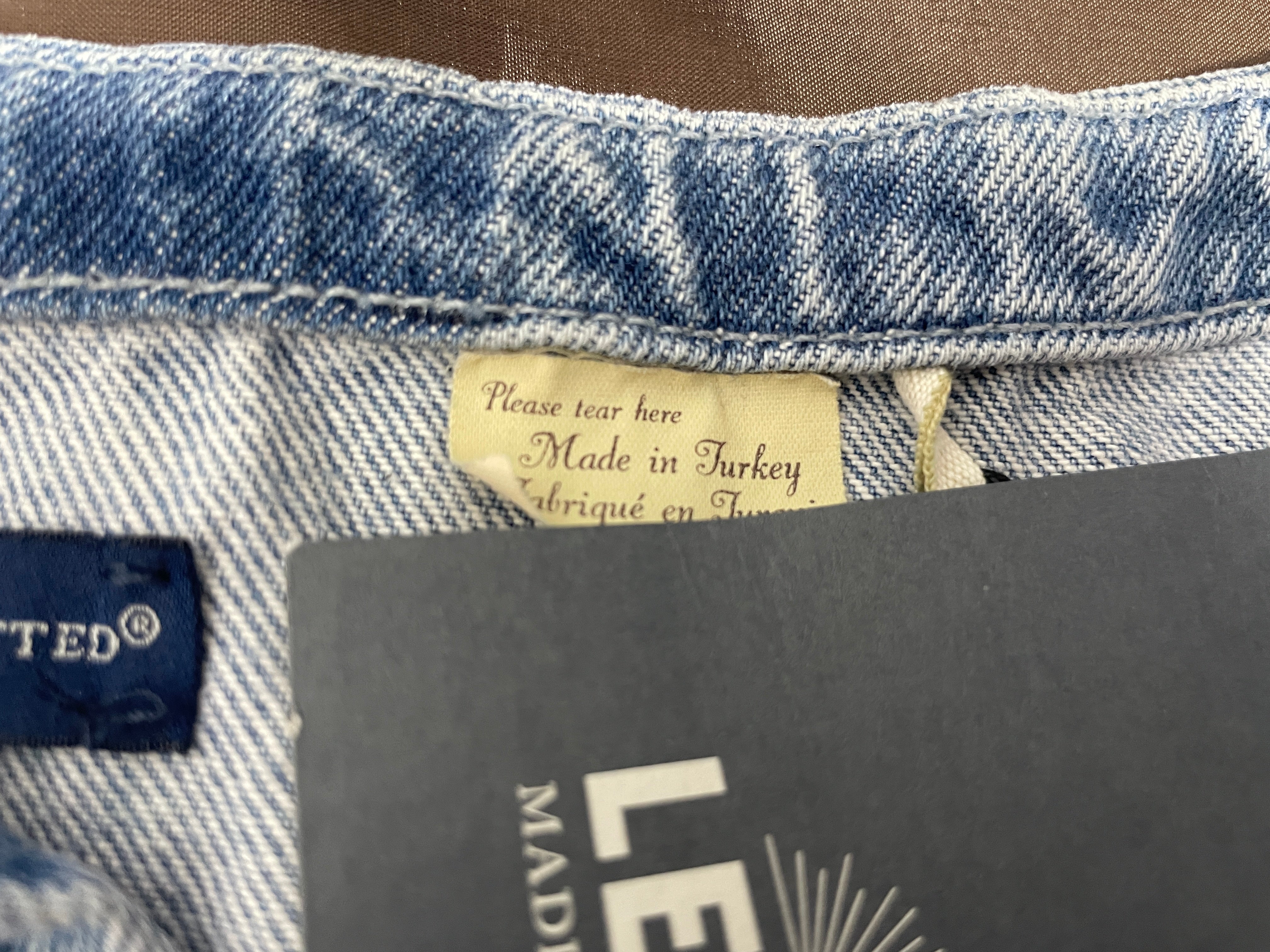 Close-up of the inside waistband of light blue denim jeans, showing a beige 'Made in Turkey' label, a blue brand tag, and a dark gray retail hangtag.