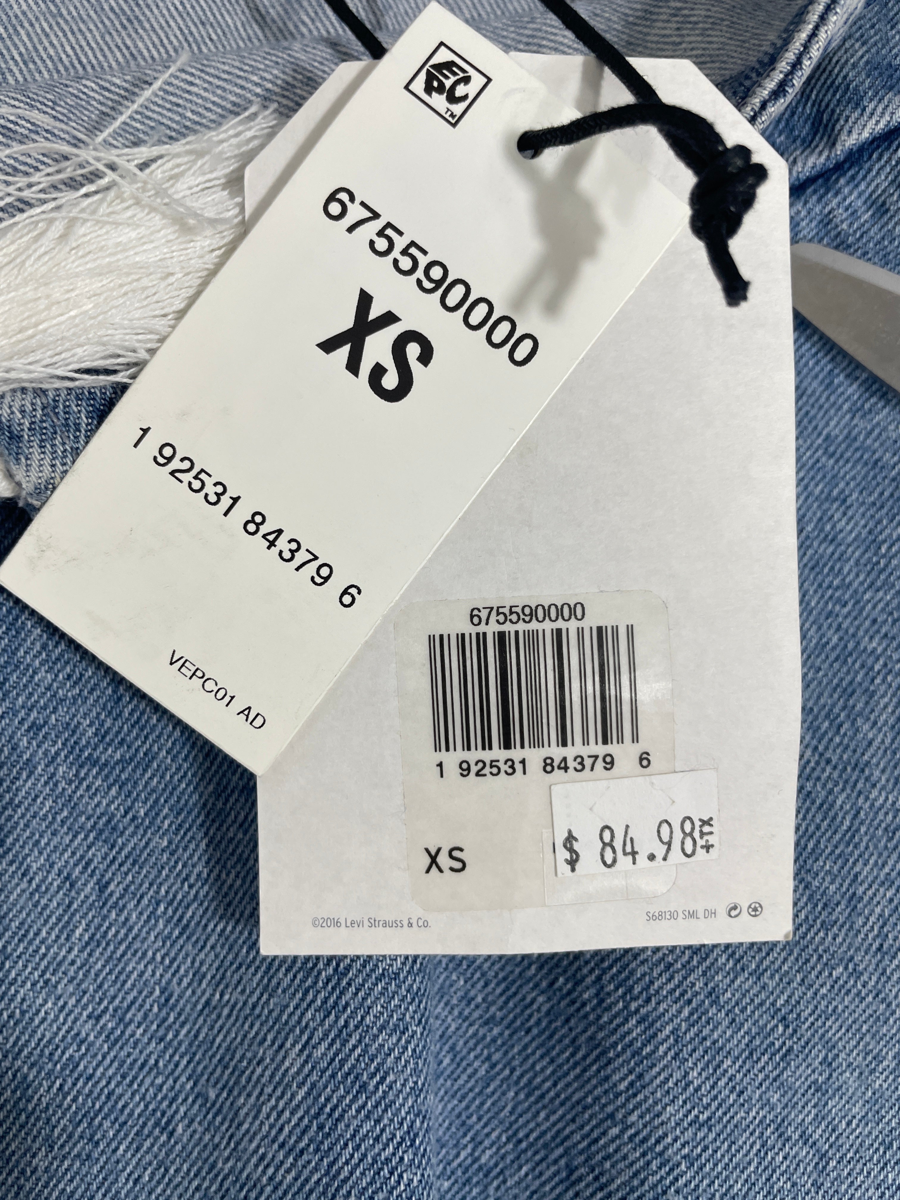 A close-up of a white garment tag attached to blue denim fabric, showing size XS, barcode, price $84.98, and a small logo near the top.