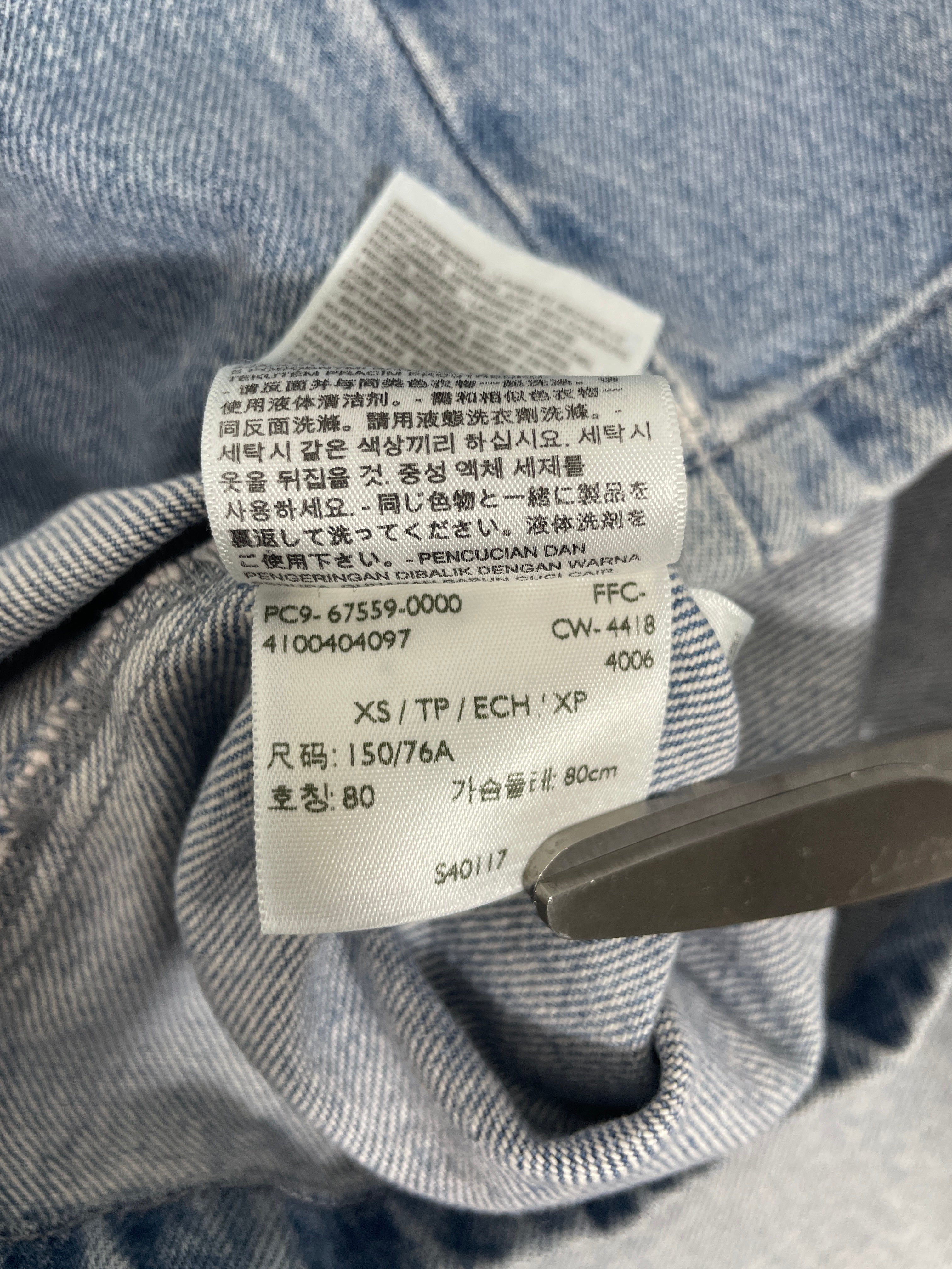 Close-up of a white fabric care label sewn onto the inside of light blue denim clothing, with a metal tool holding the label in place.