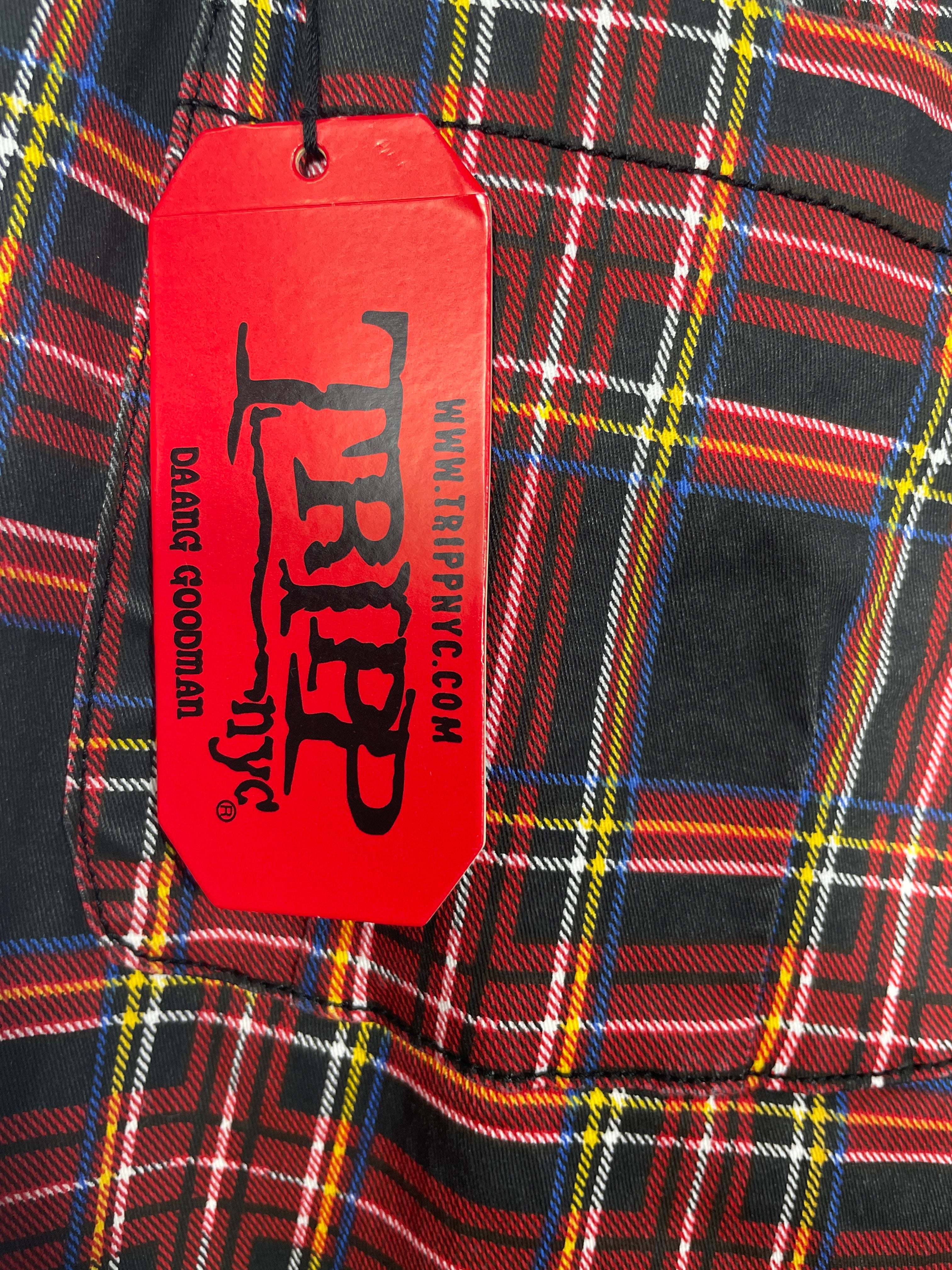 A close-up of a red plaid garment with a bright red Tripp brand hangtag attached. The plaid pattern features black, red, yellow, and white stripes.