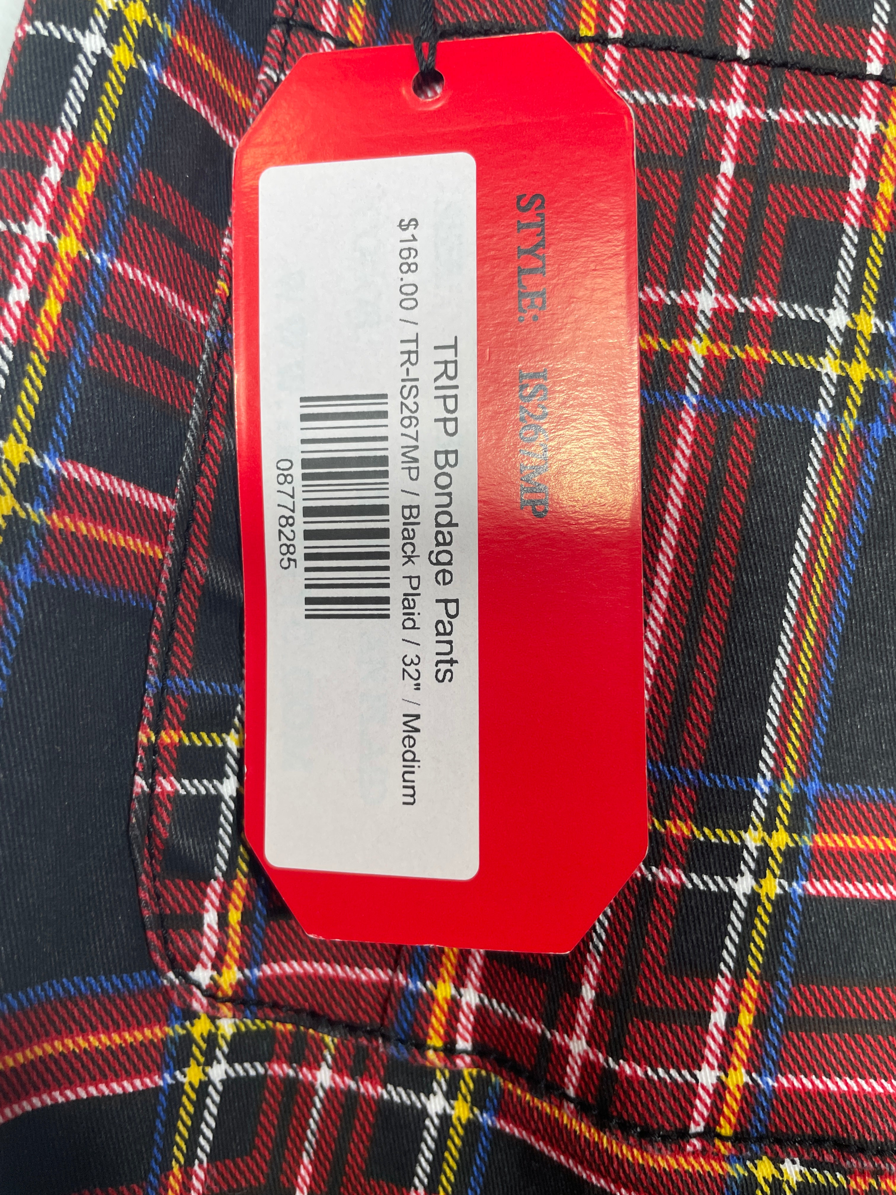 A close-up of a red plaid garment tag attached to a red price label, with the text 'STYLE: STUNNER' and 'TRIP Binding Pants' visible. The fabric shows a multicolor plaid pattern with red, yellow, black, and white.