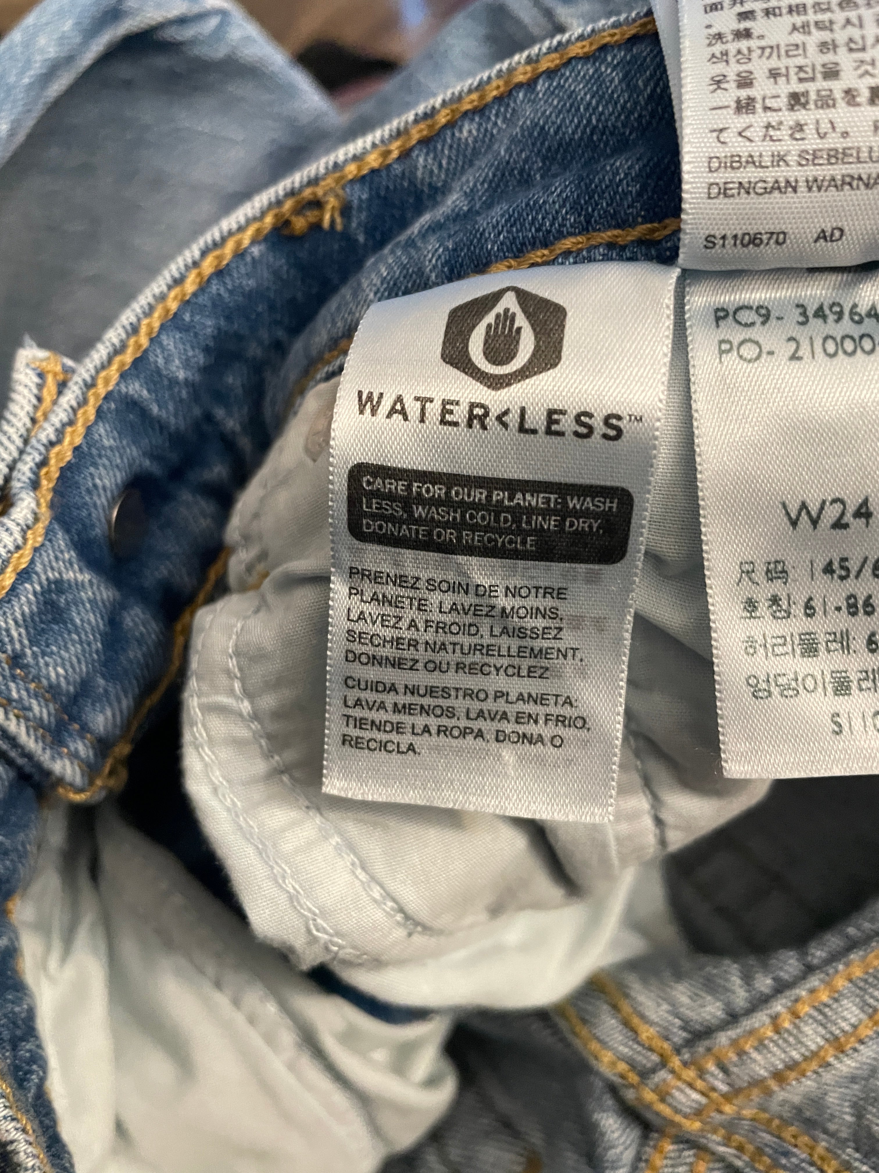 Close-up of a denim garment showing the inner label area, with a prominent Waterless care label and other sewn-in tags visible.