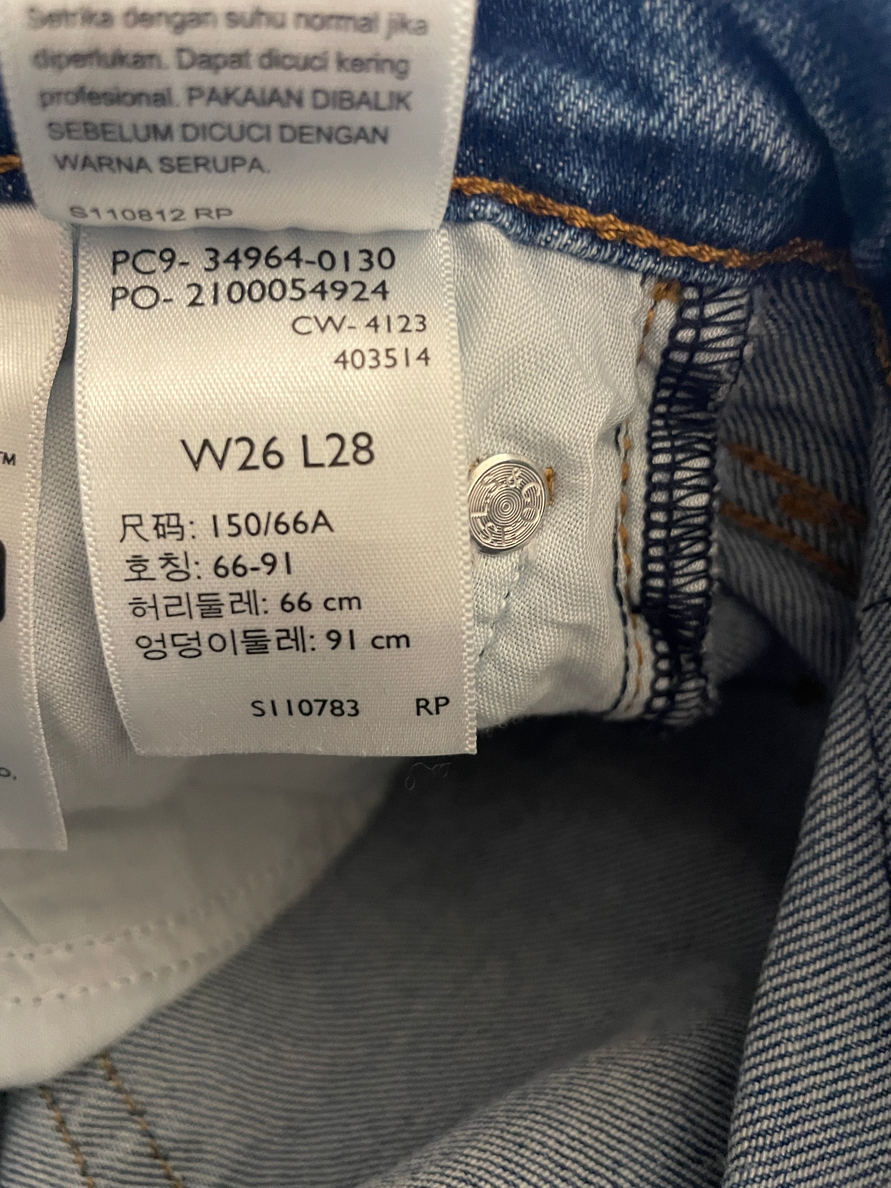 Close-up of a clothing size label sewn onto the inside of a blue denim jacket, showing garment code W26 L28 and size information in Indonesian and Korean.