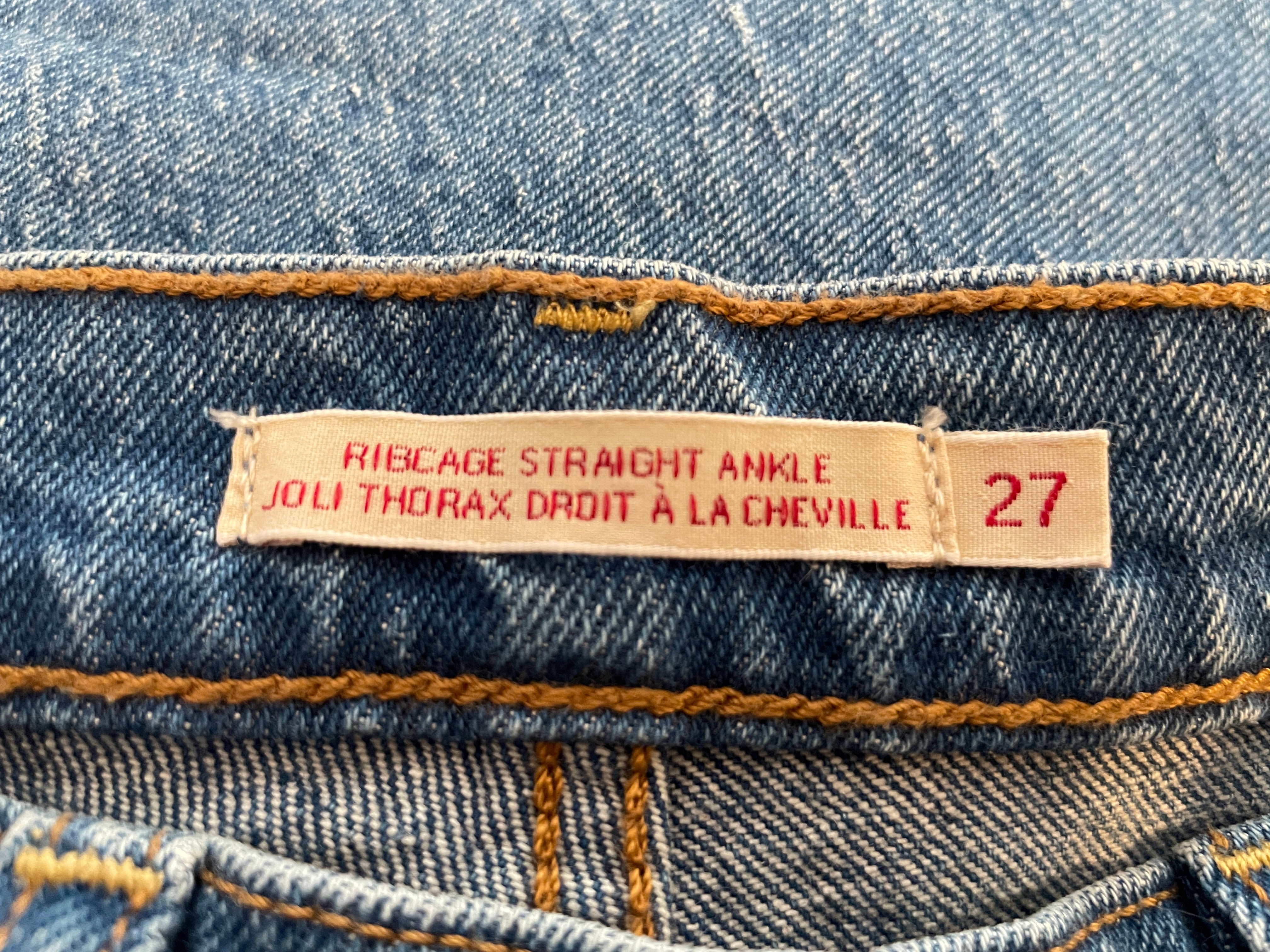 Close-up of a denim waistband label reading 'RIB CAGE STRAIGHT ANGLE JOLI THORAX DROIT A LA CHEVILLE 27' on blue denim fabric with orange stitching.