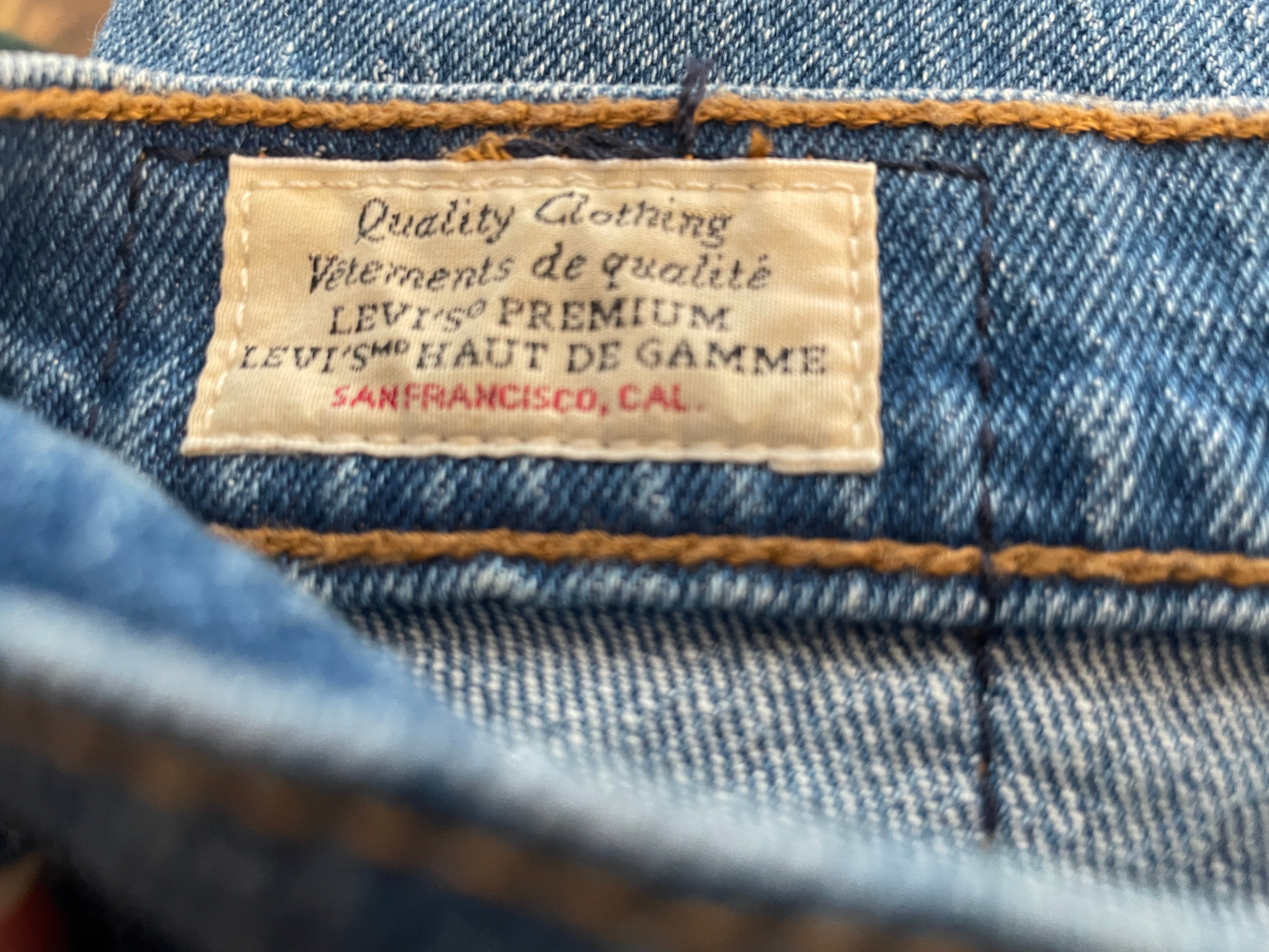 Close-up of a denim garment showing a sewn-in label that reads 'Quality Clothing' and 'Vieillissements de qualité Levi's Premium', with blue denim fabric and orange-brown contrast stitching visible.