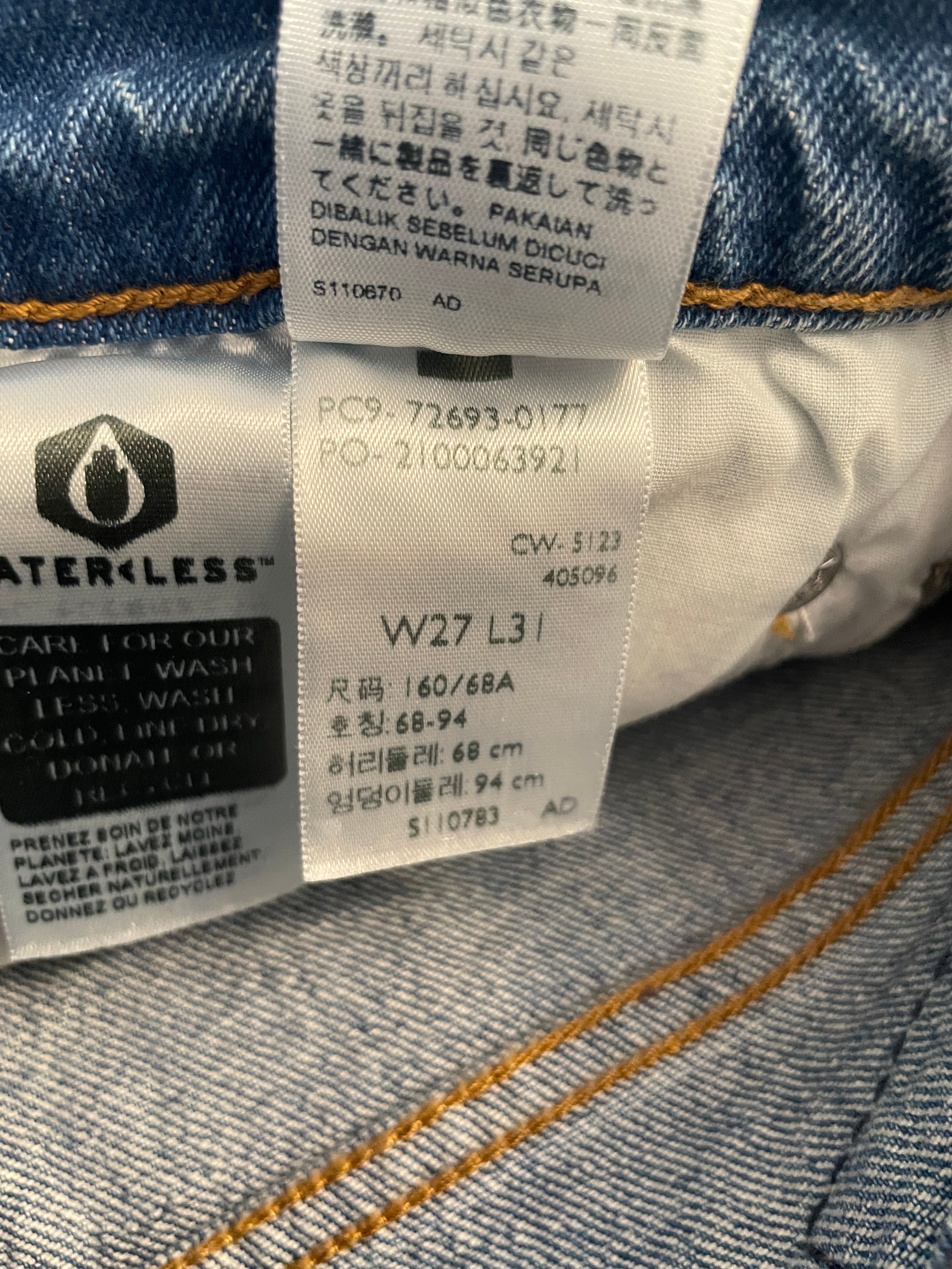 Close-up of a denim garment's interior labels, including a Waterless care label and a size tag reading W27 L31, with blue denim fabric and orange contrast stitching visible.