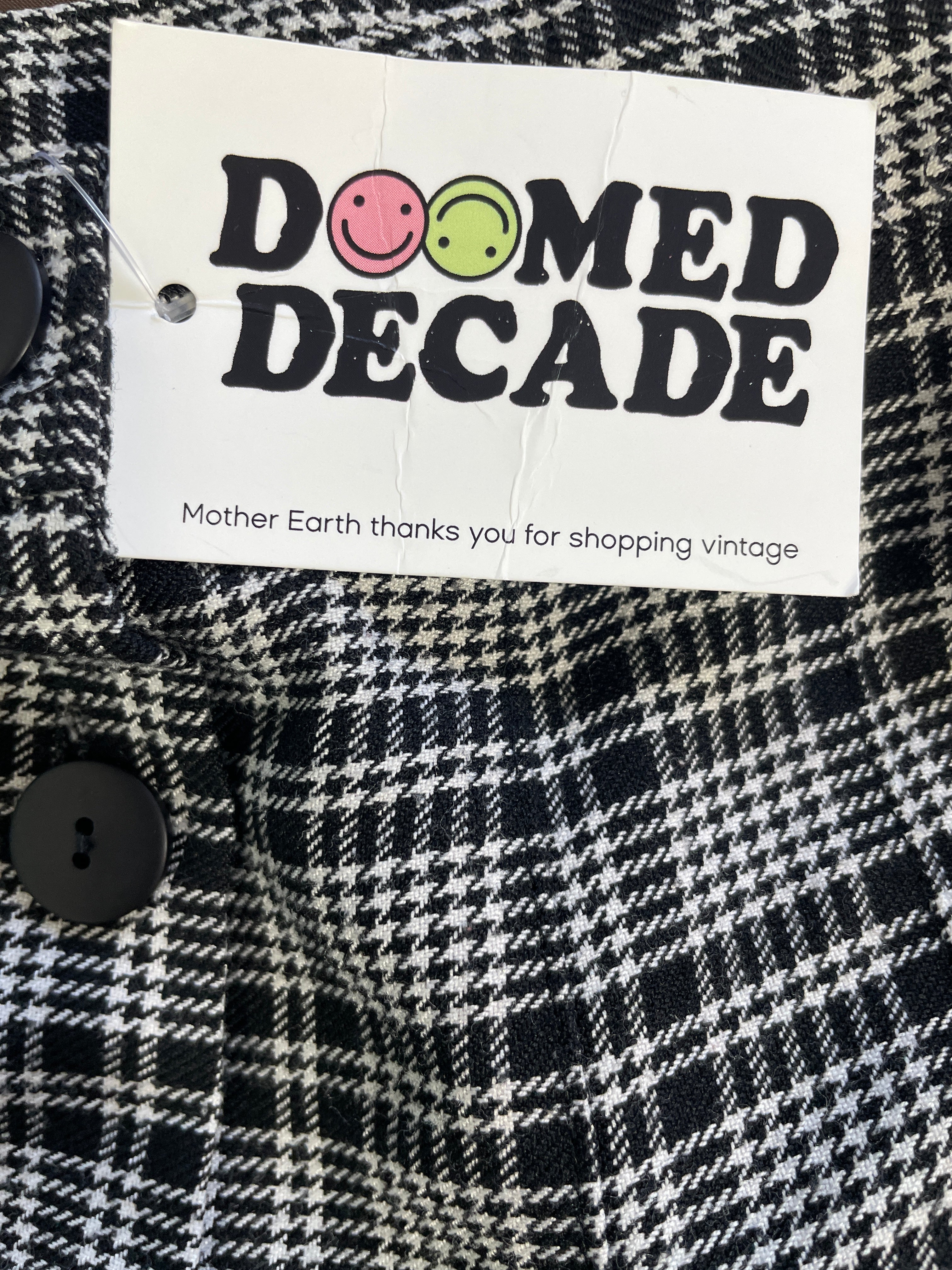 A close-up of a black and white plaid garment with a white paper hangtag reading 'DOomed Decade' and 'Mother Earth thanks you for shopping vintage.'.