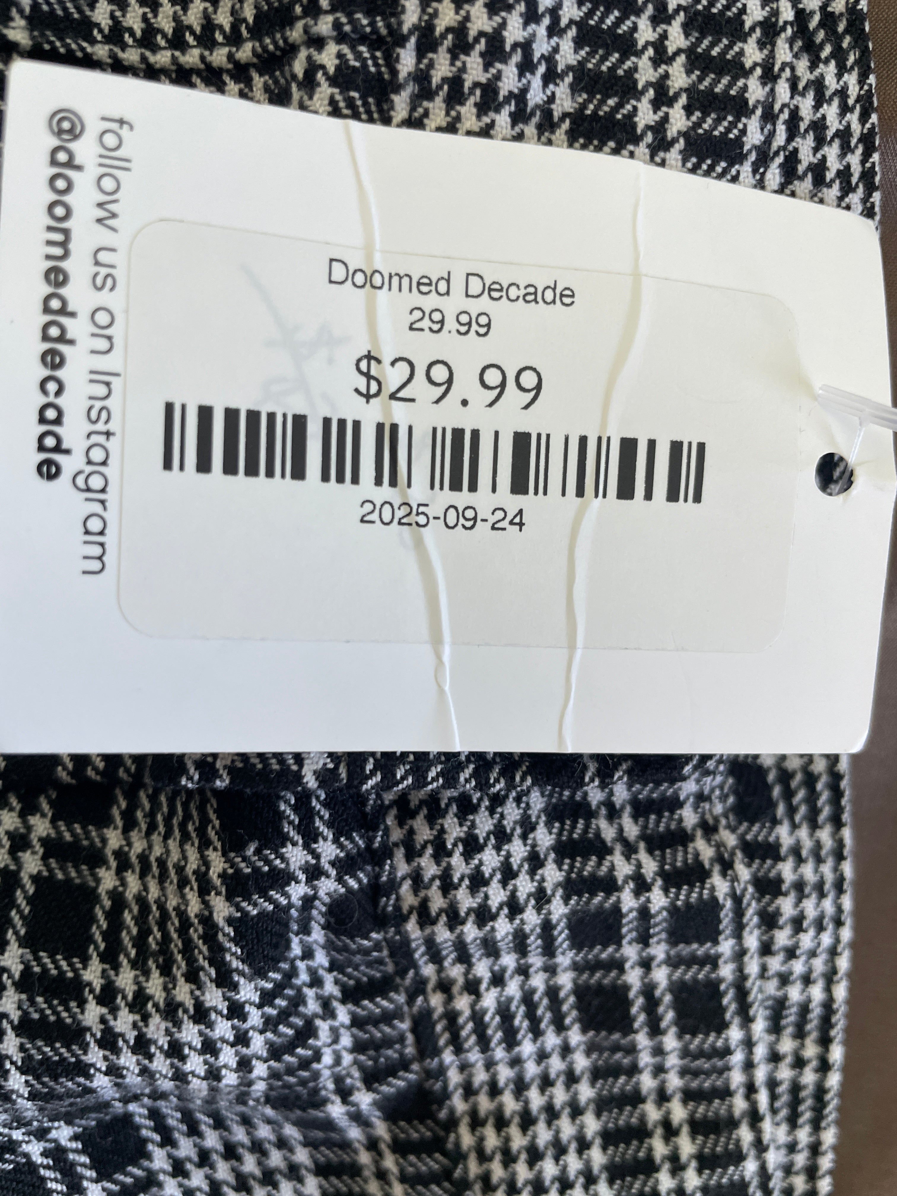 A close-up of a black and white houndstooth patterned fabric with a white paper price tag attached. The tag reads 'Doomed Decade', '$29.