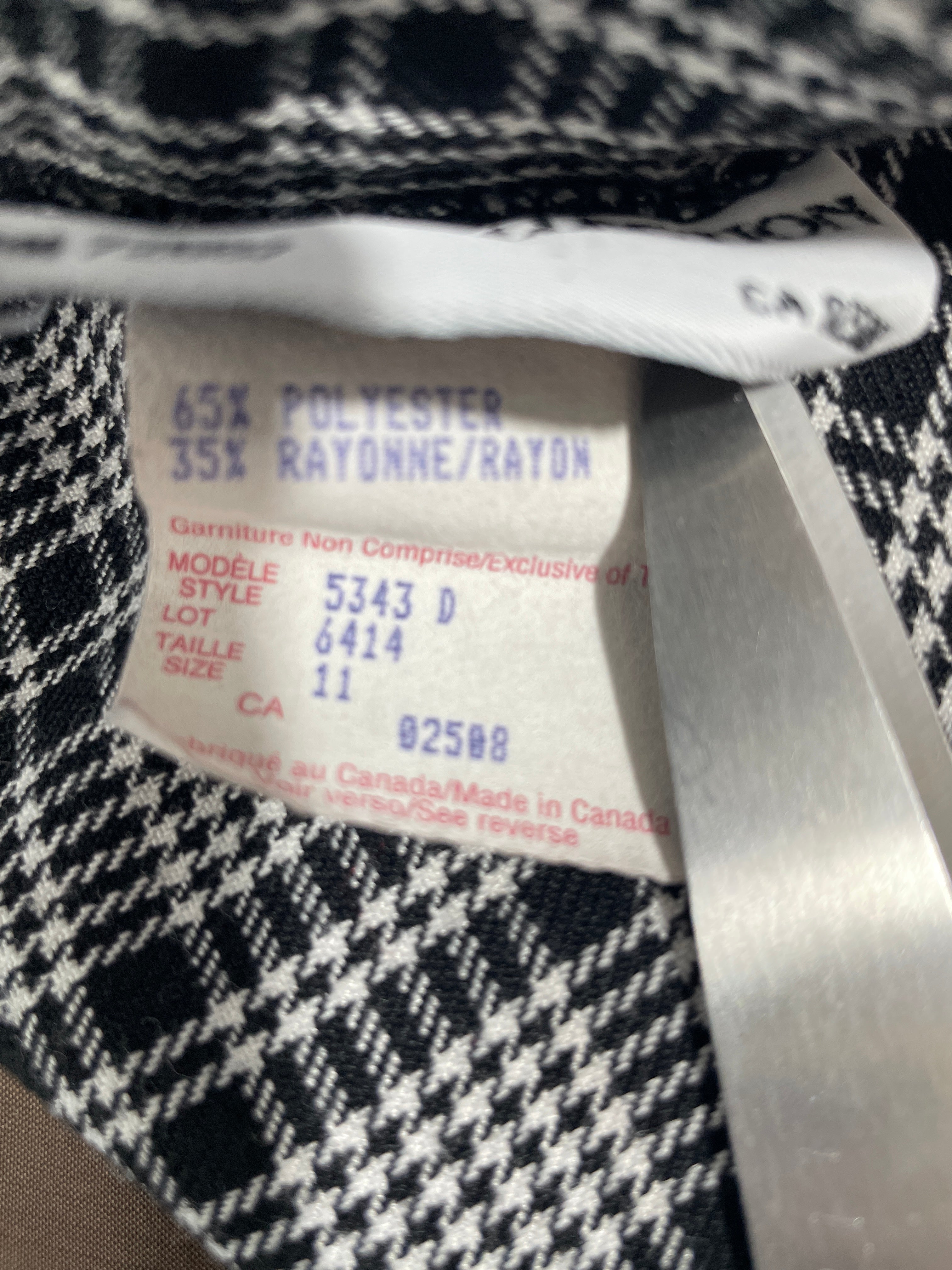 Close-up of a garment's interior size and care label sewn onto a black and white plaid fabric, showing text including '65% POLYESTER 35% RAYONNE/RAYON'.