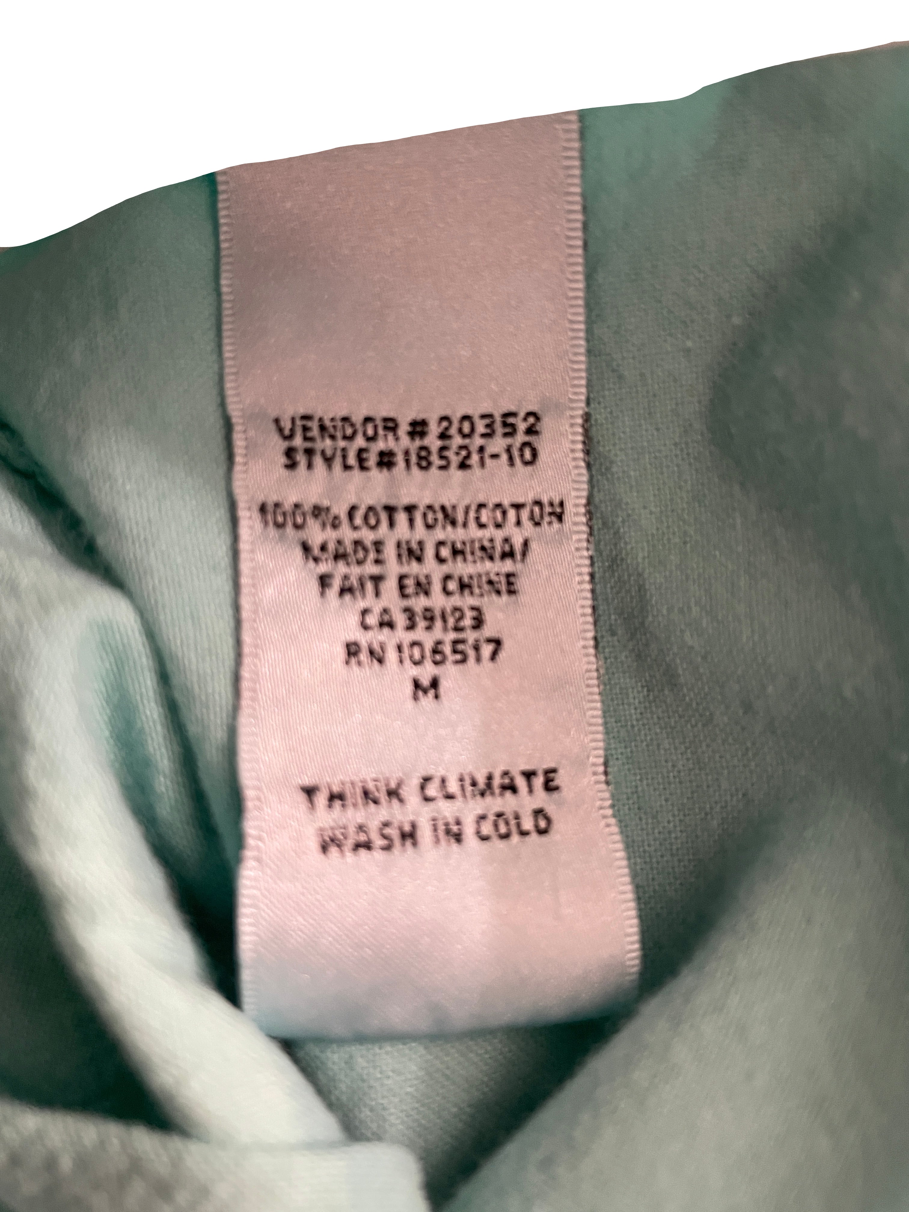 Close-up of a garment's interior neck label sewn onto light sage green fabric. The label reads 'VENDOR # 20352 STYLE#18521-10 100% COTTON/COTON MADE IN CHINA/FAIT EN CHINE CA 39123 RN 106517 M THINK CLIMATE WASH IN COLD'.