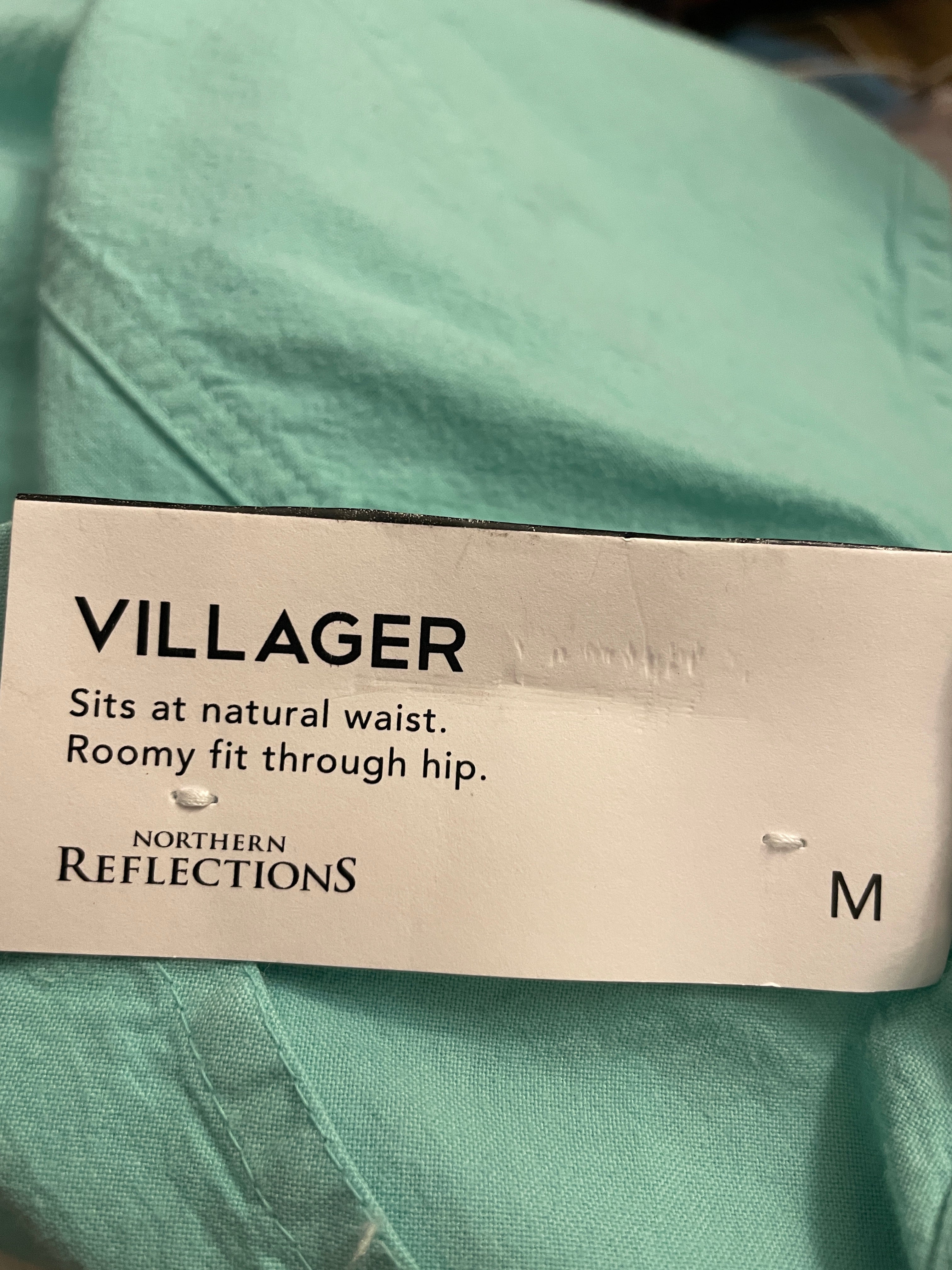A close-up of a mint green garment tag reading 'VILLAGER' with fit details and the brand 'NORTHERN REFLECTIONS', placed on folded matching fabric.
