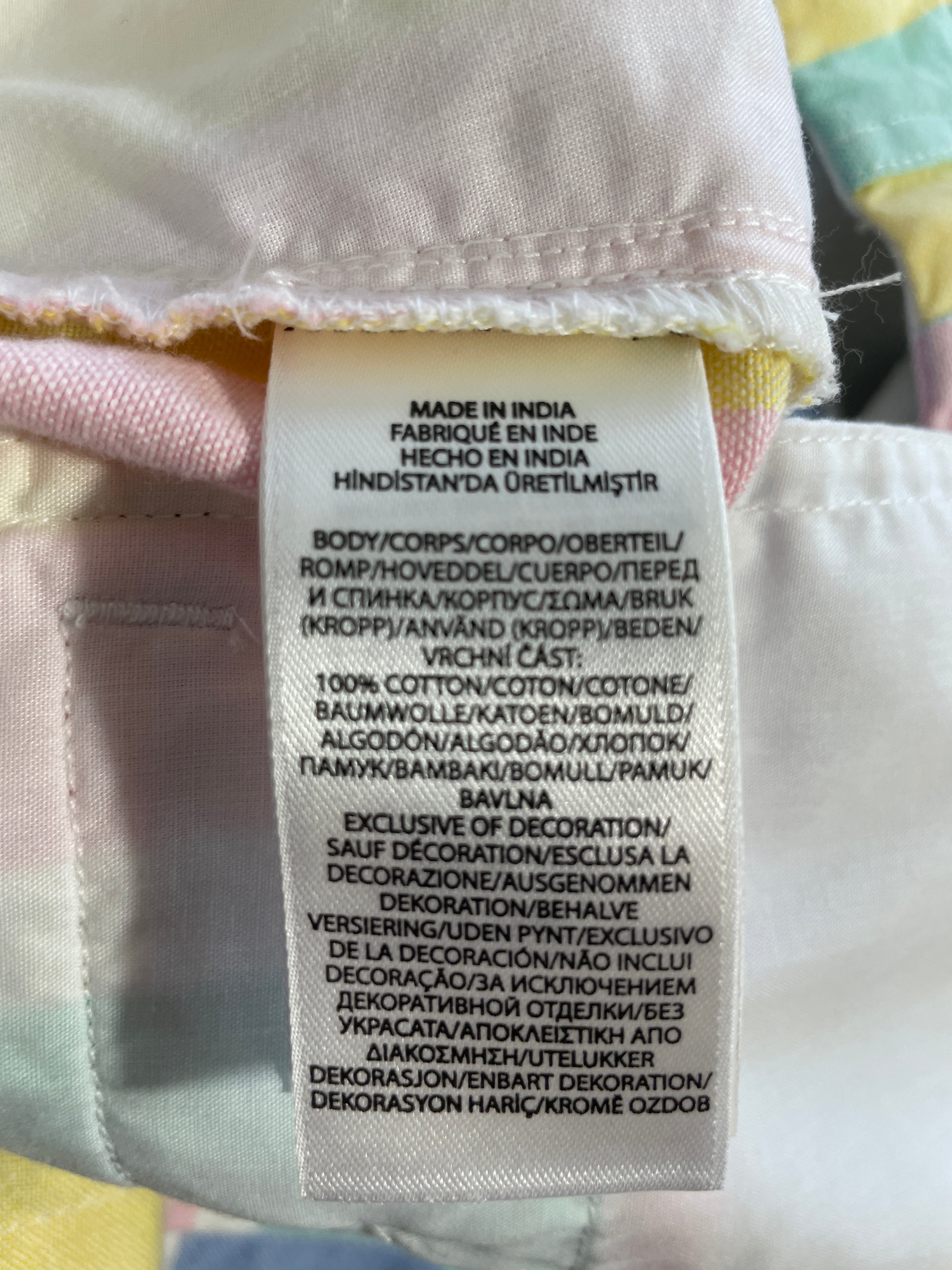 A close-up of a white fabric care label sewn onto a light pink garment. The label contains printed text in multiple languages stating 'MADE IN INDIA' and describing the product as 100% cotton/coton.