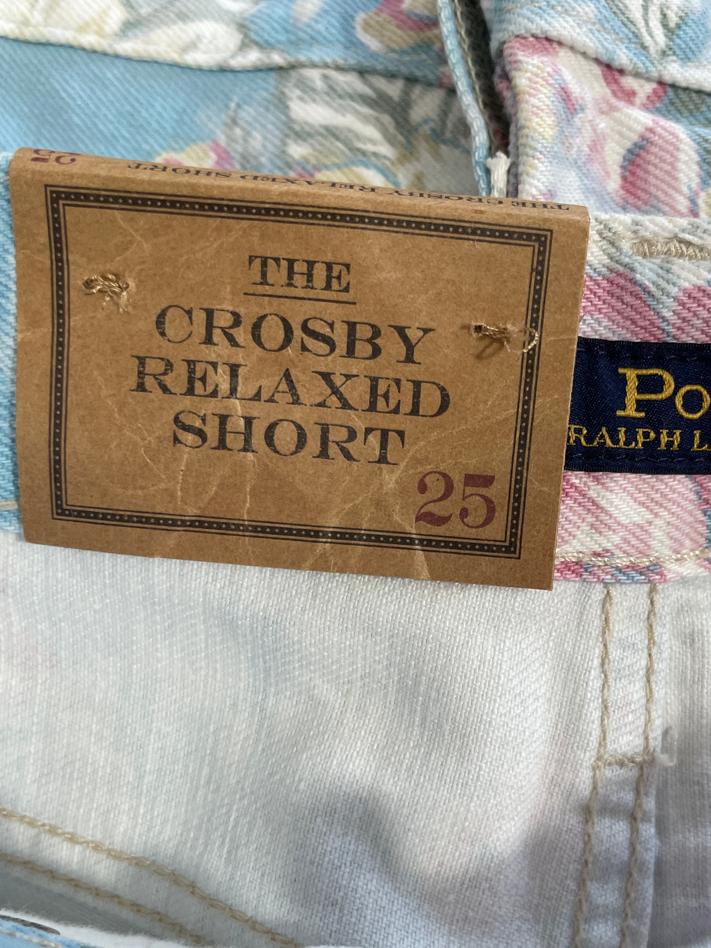 Close-up of Polo Ralph Lauren shorts with a brown paper tag reading 'THE CROSBY RELAXED SHORT 25' and a navy Polo Ralph Lauren brand label, showing the floral-patterned fabric and white washed denim waistband.