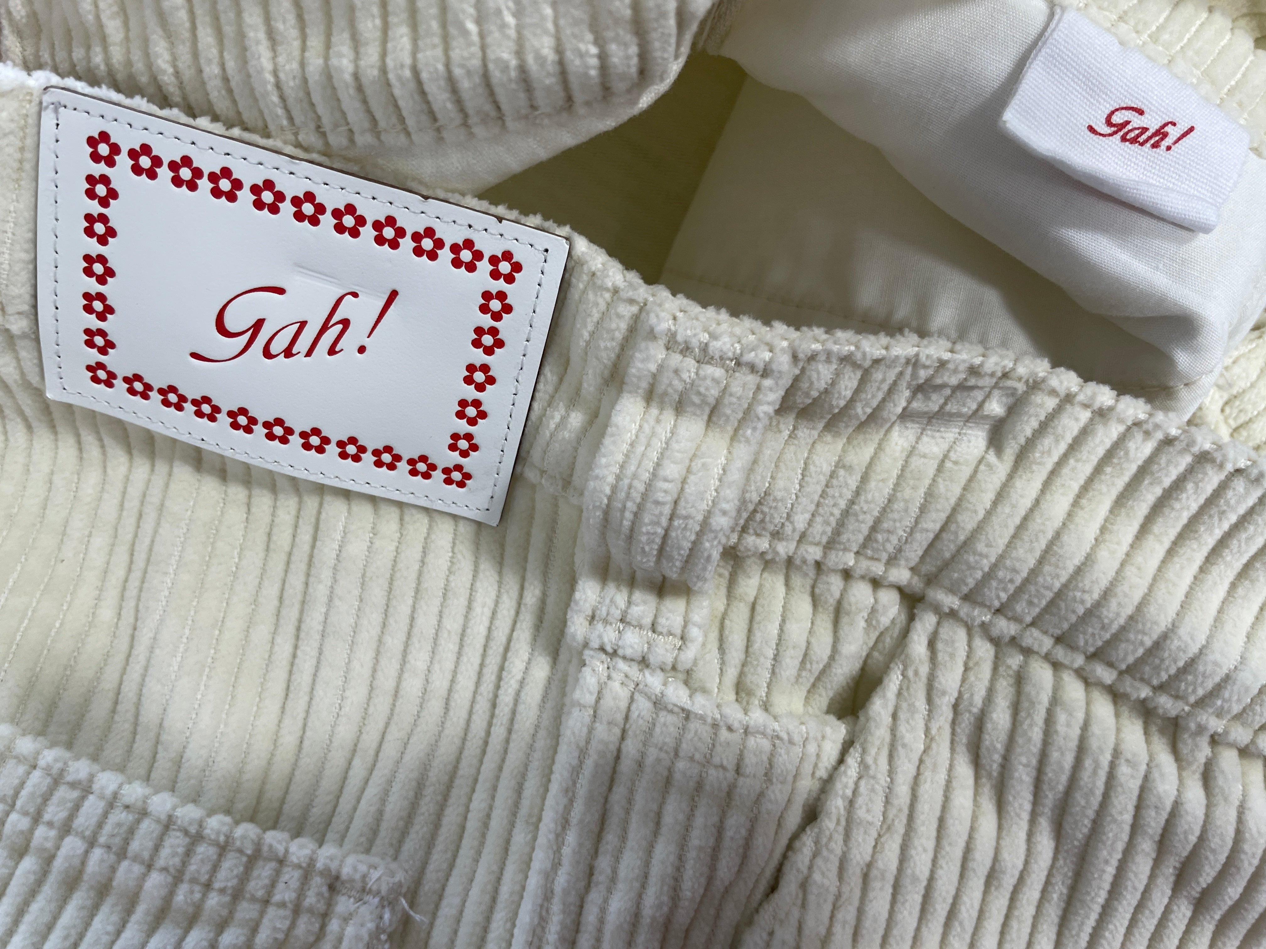 Close-up of a cream-colored ribbed corduroy garment, likely a baby jacket or romper, featuring a white brand label with a red floral border and the text 'Gah!' on the inside neck.