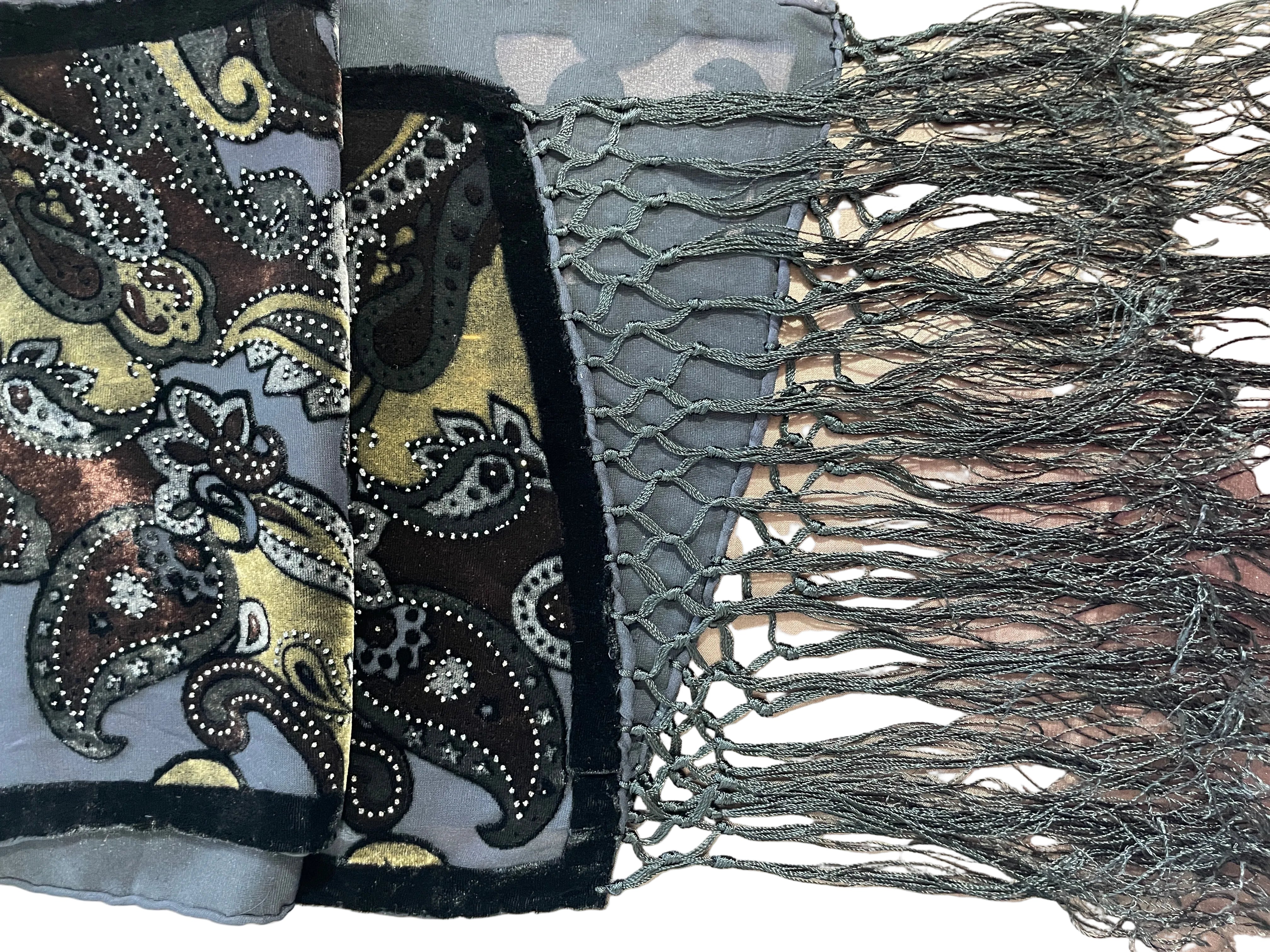 A close-up of a scarf featuring a mixed-media design with a paisley pattern on one side, a gray camouflage pattern on another, and a dark gray knotted fringe on the other side.
