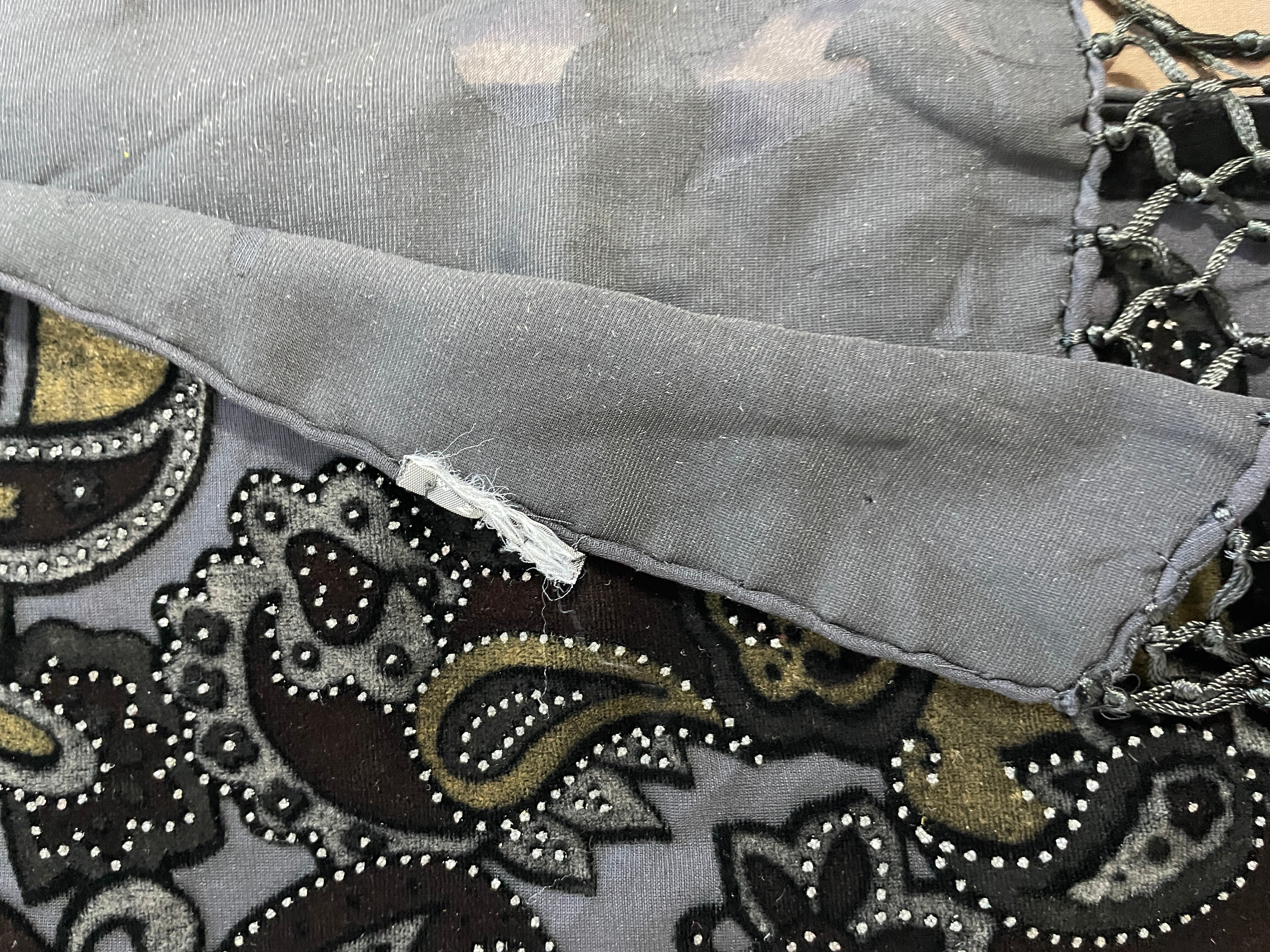 Close-up of a gray fabric textile with a paisley pattern in black, brown, and gold, featuring decorative beadwork and a small white thread detail.