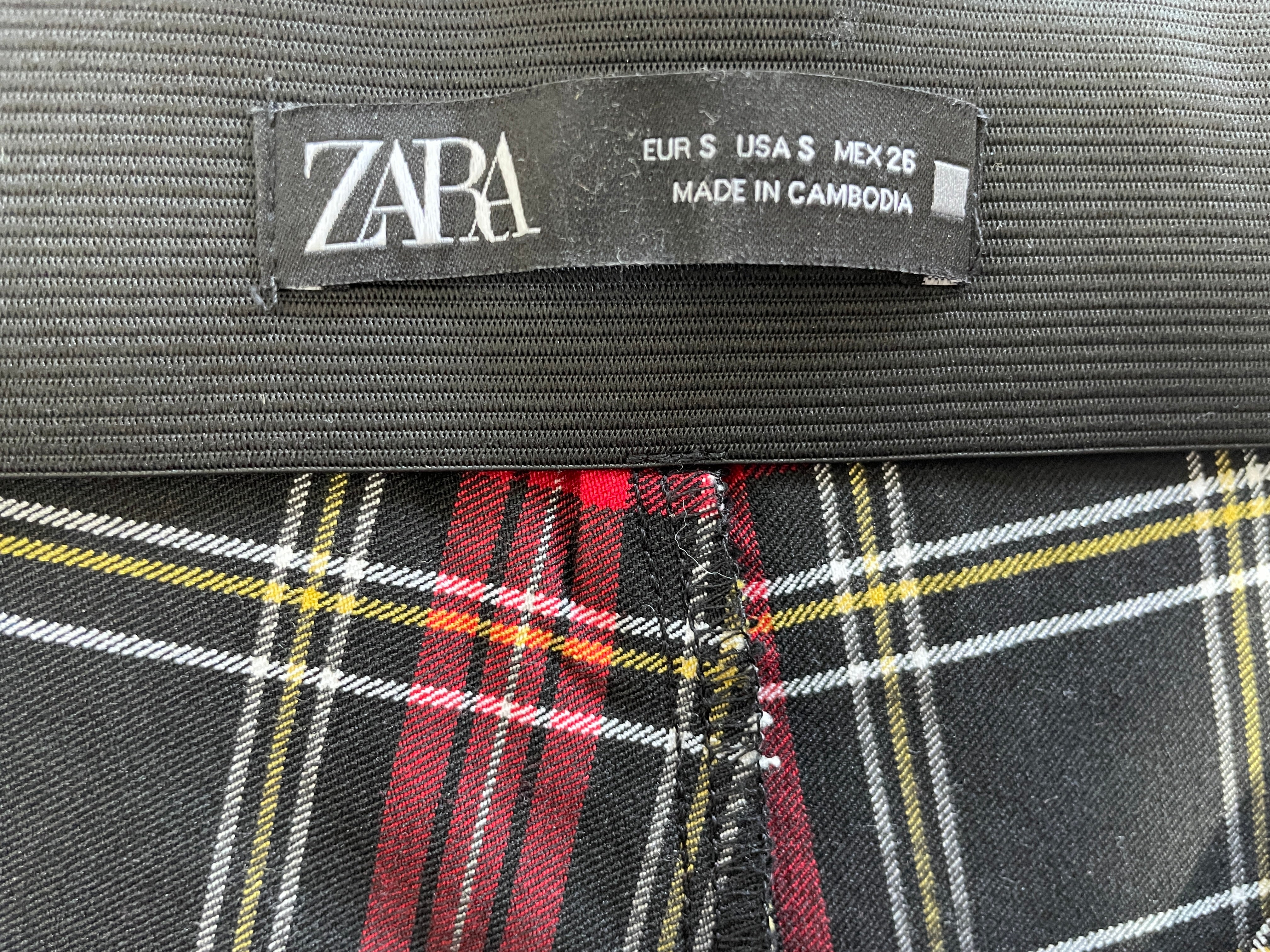 Close-up of a ZARA garment label sewn onto a plaid fabric, showing the brand name and size information.