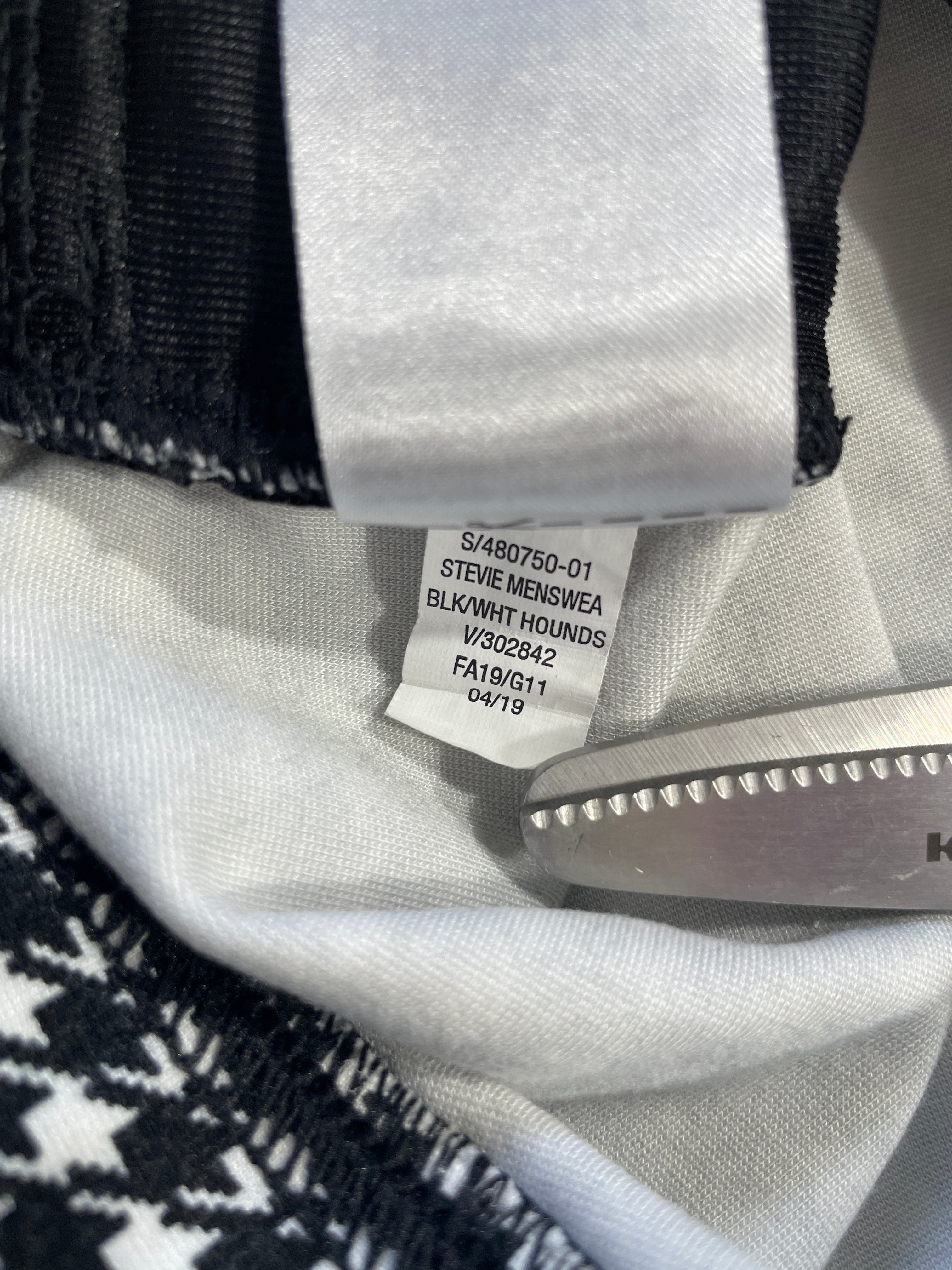 Close-up of a garment's interior neck label reading Steve Meinswea Blk/White Hounds, with a metal tag inserted into the collar.