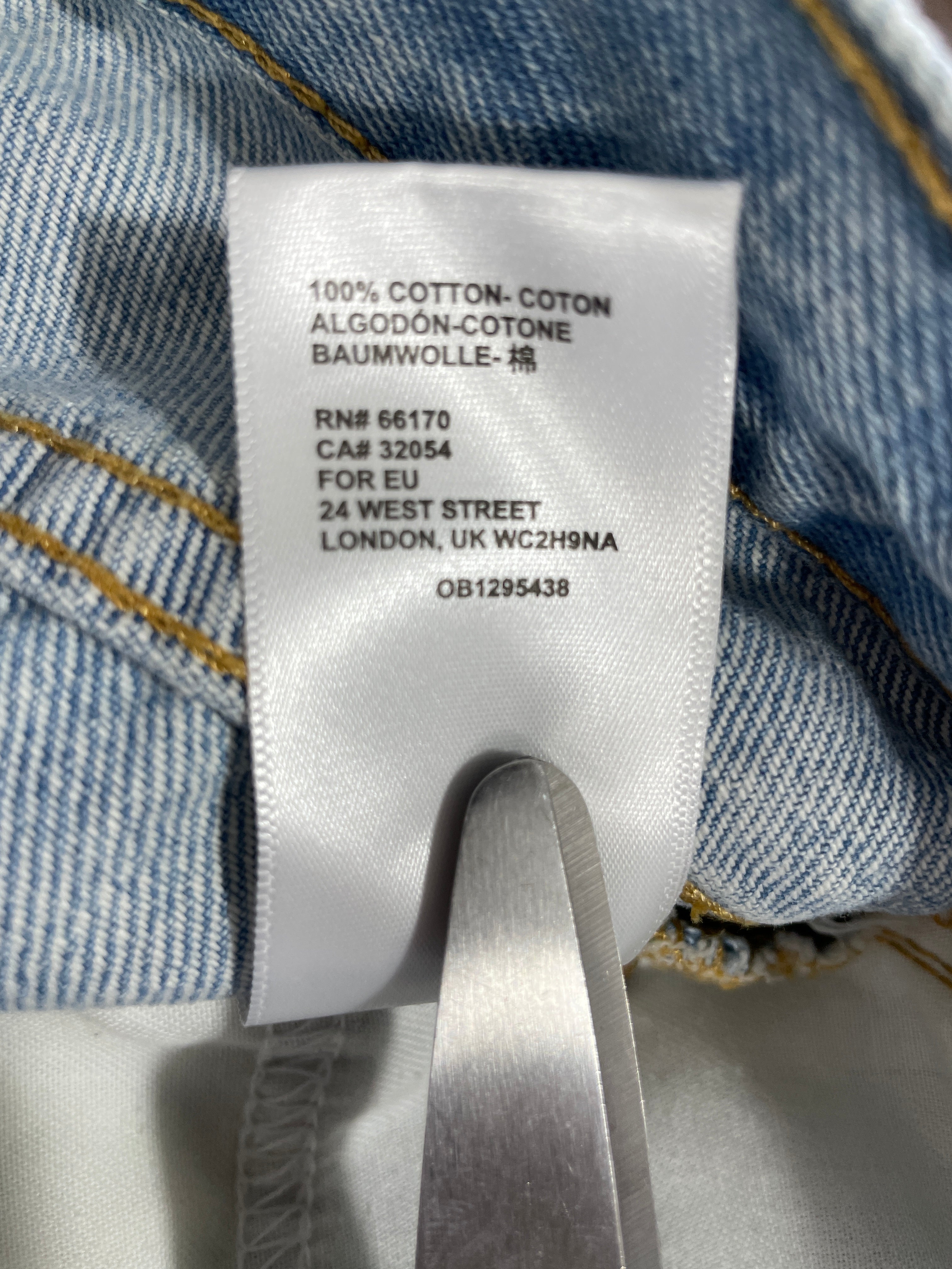 Close-up of a white fabric care label sewn onto a light blue denim garment, with a metal tool holding the label in place. The label reads '100% COTTON- COTON ALGODÓN-COTONE BAUMWOLLE-棉' and includes manufacturer text for London.
