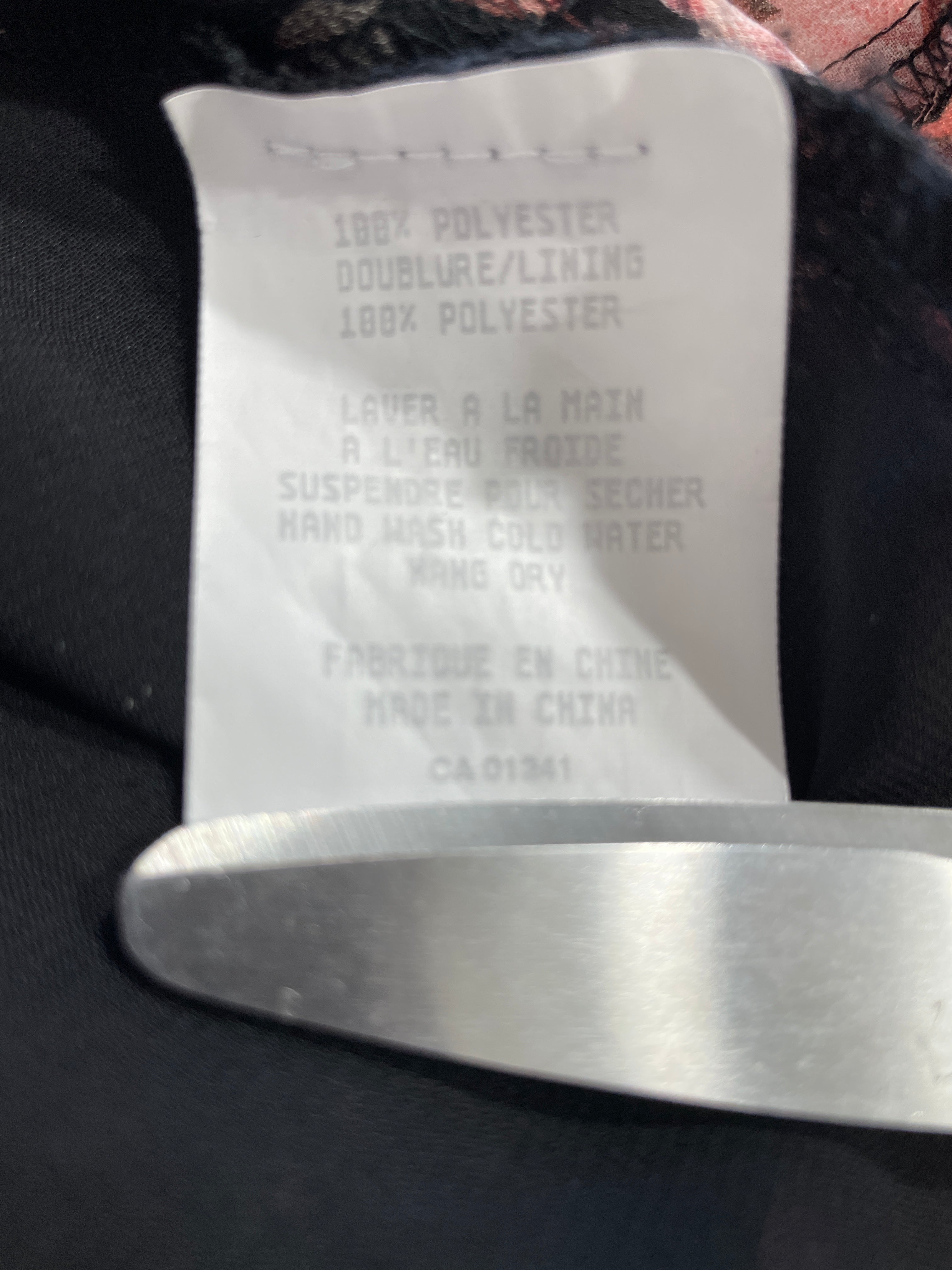 Close-up of a white sewn-in size tag on black fabric, showing text including '100% POLYESTER' and 'MADE IN CHINA', with a silver metal edge visible at the bottom.